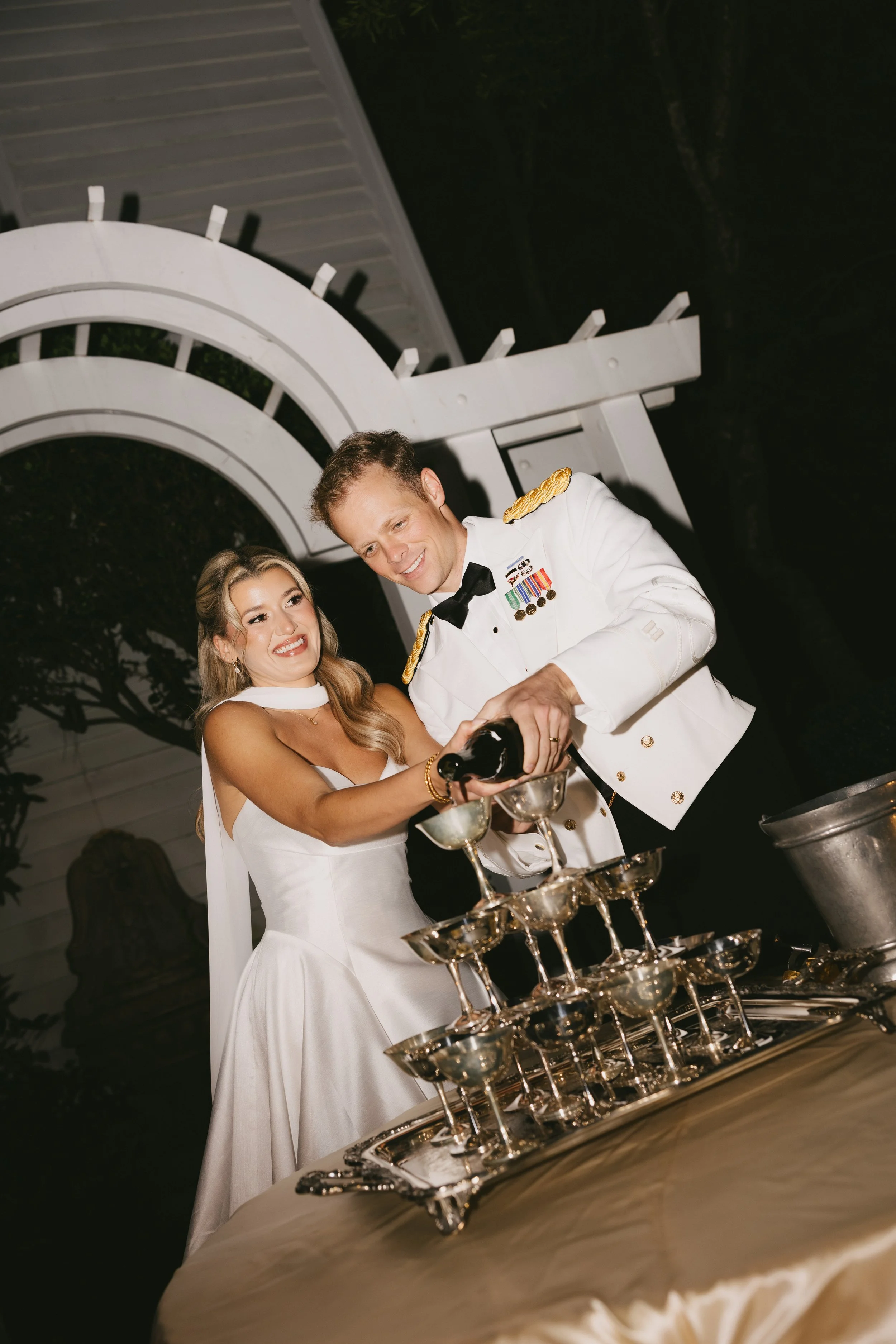 A bride and groom pouring champagne into glasses at their wedding celebration. The bride is in a white wedding dress with a veil, smiling at the groom, who is in a white military uniform with medals, also smiling.