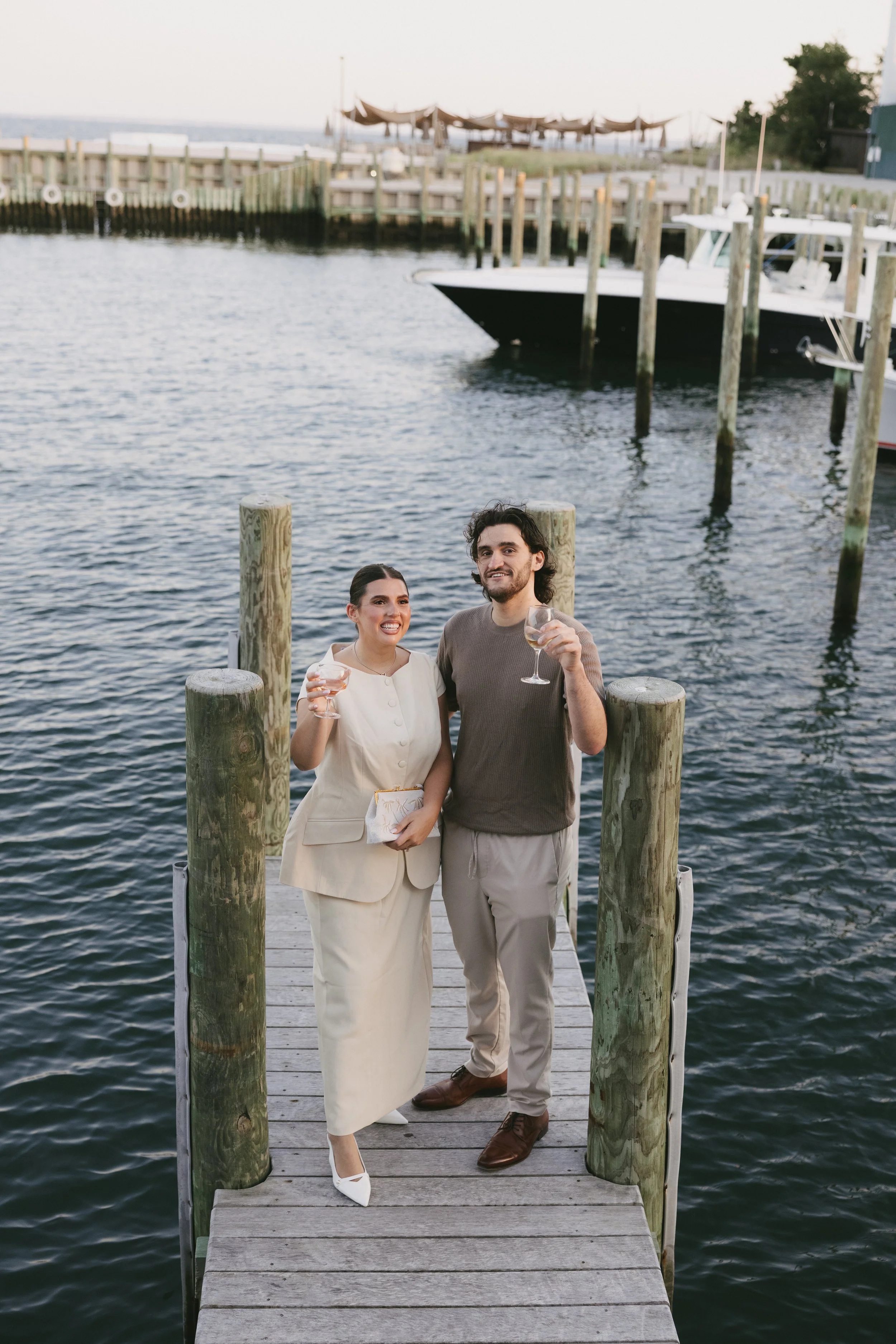 A couple holding wine glasses on a boat dock, smiling with a marina of boats and water in the background.