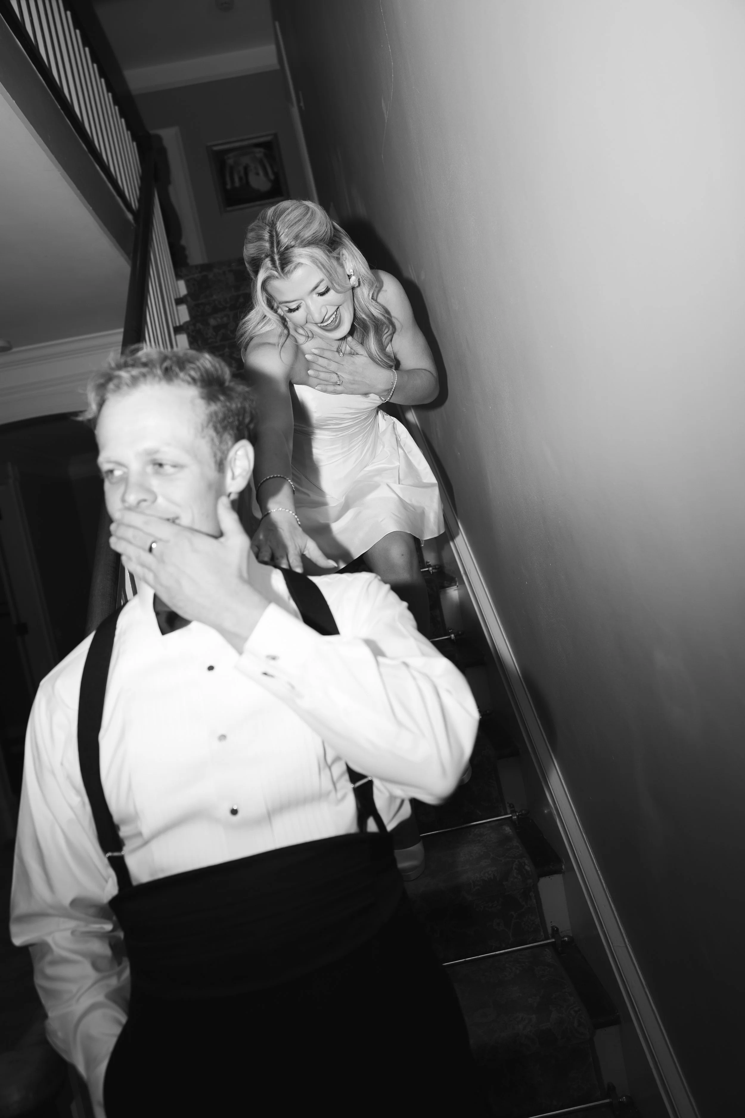 Black and white photo of a woman in a wedding dress and a man in formal attire on a staircase, both appear surprised or amused.