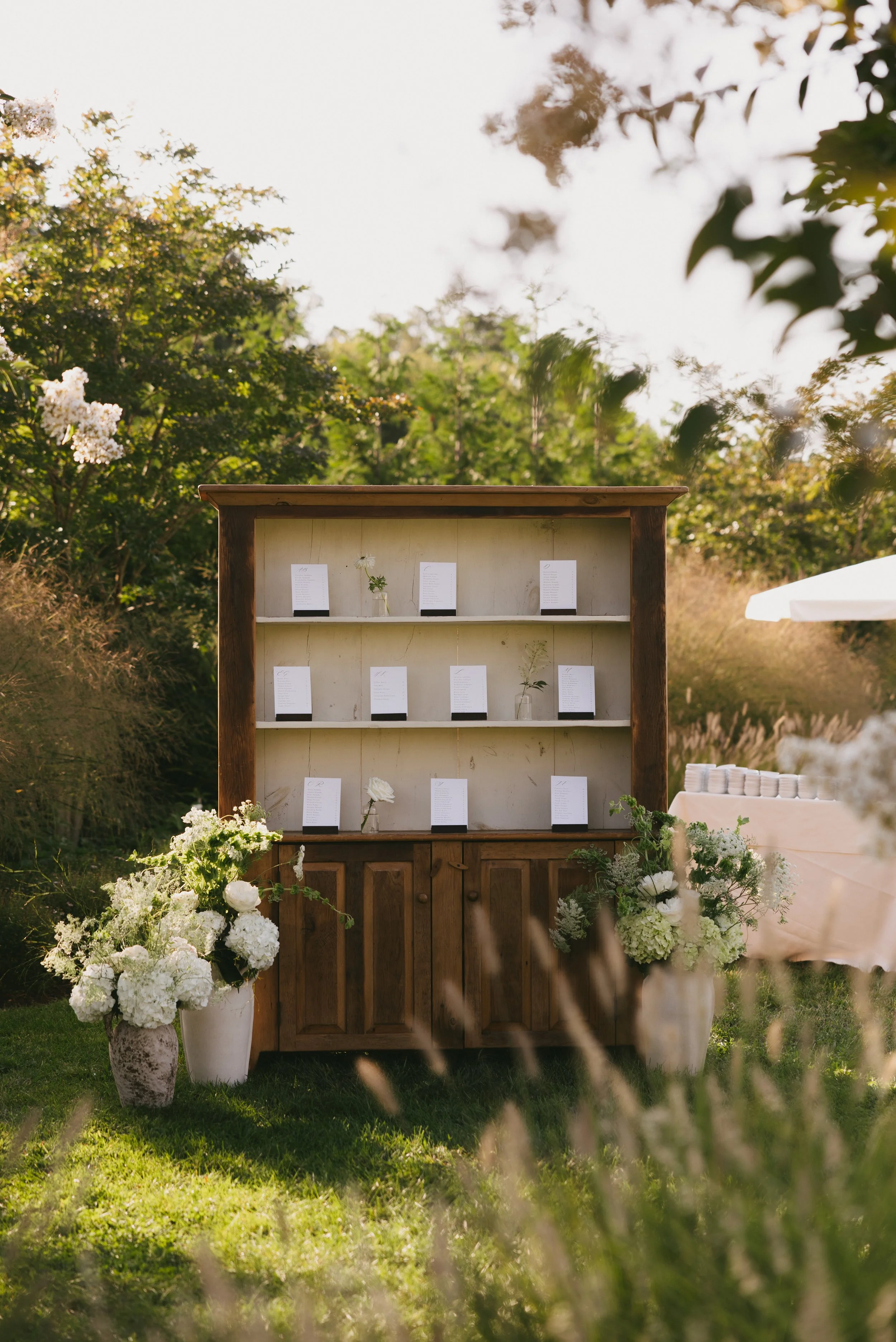 Rustic outdoor wedding or event seating chart display with white flowers in vases, set in a garden with trees and greenery.