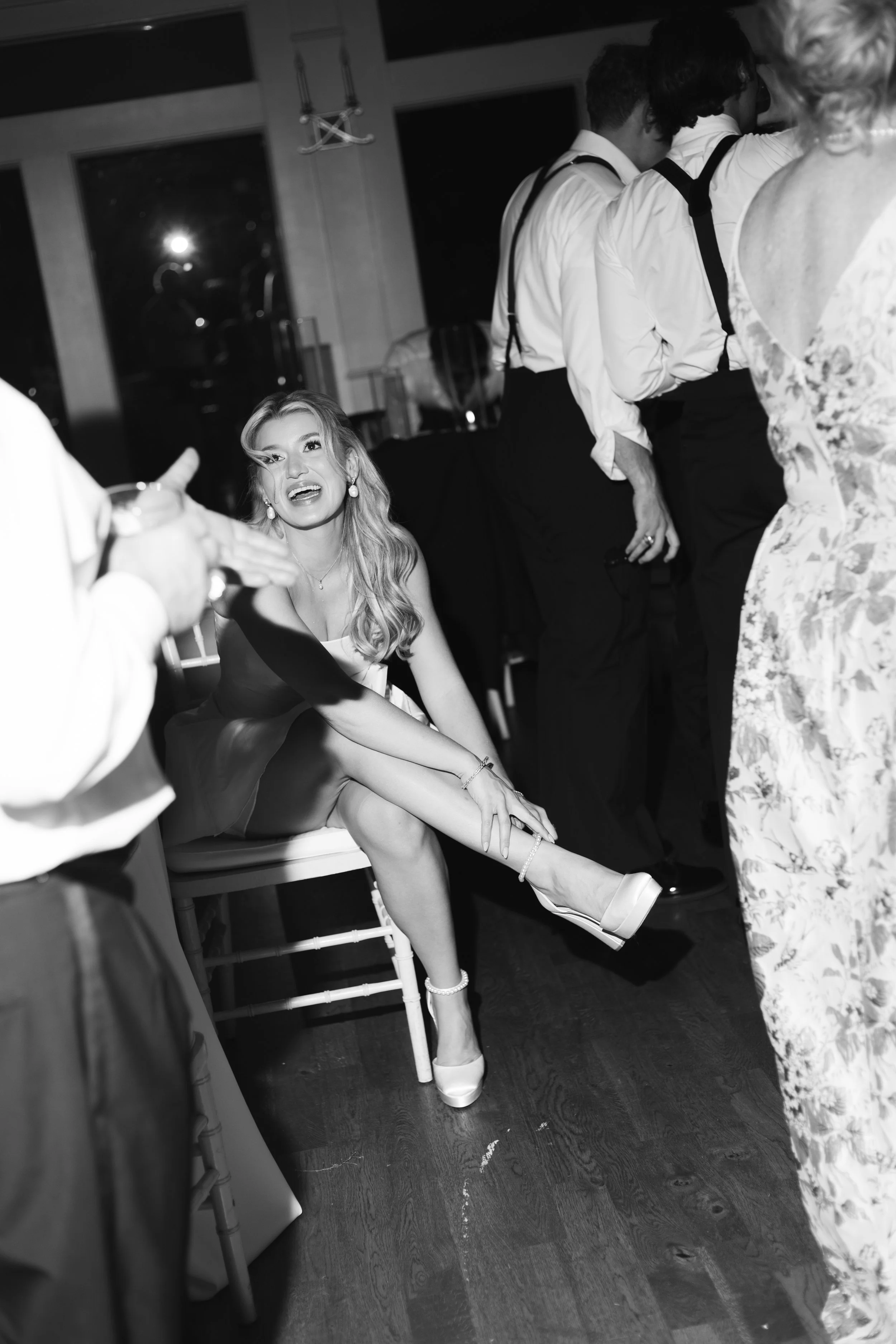 A woman with long blonde hair is sitting on a chair at a social event, wearing a light-colored dress and high heels, and appears to be speaking or laughing while interacting with others around her.