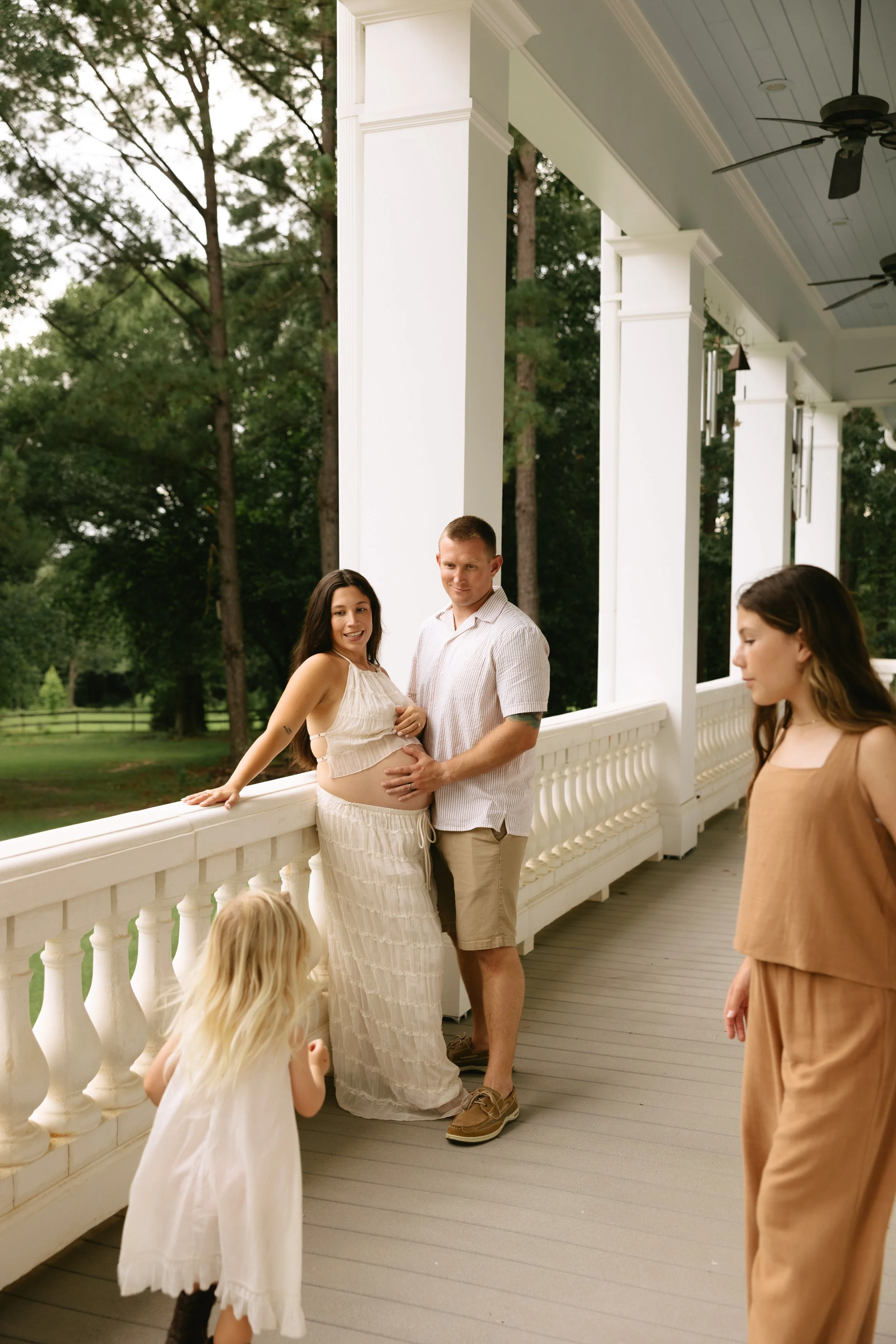 A pregnant woman and her partner posing on a porch with two young girls nearby.