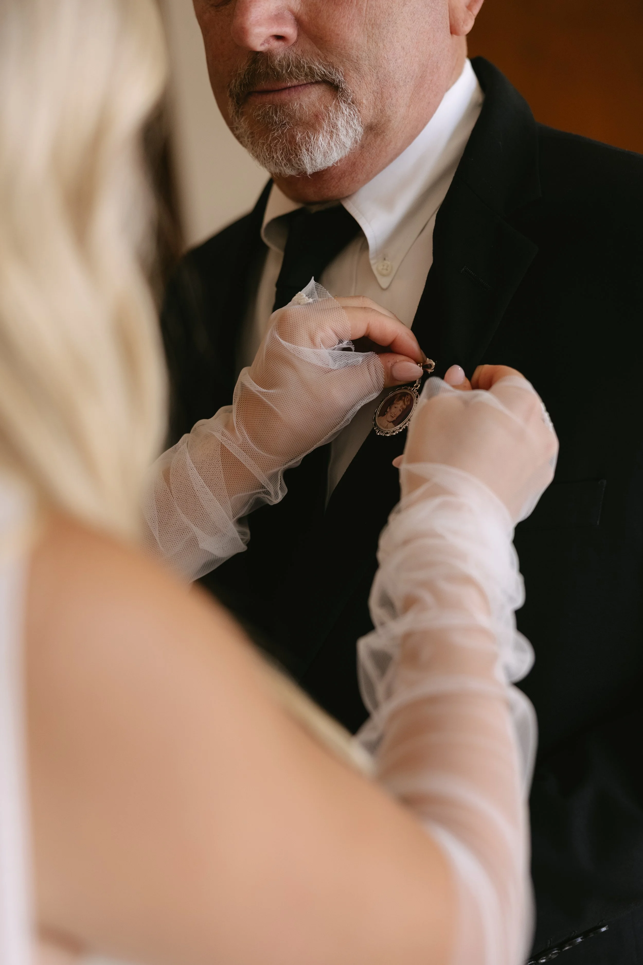 A woman in wedding dress with lace sleeves pins a wedding medal onto a man's black tuxedo. The man has a gray beard and is in an indoor setting.