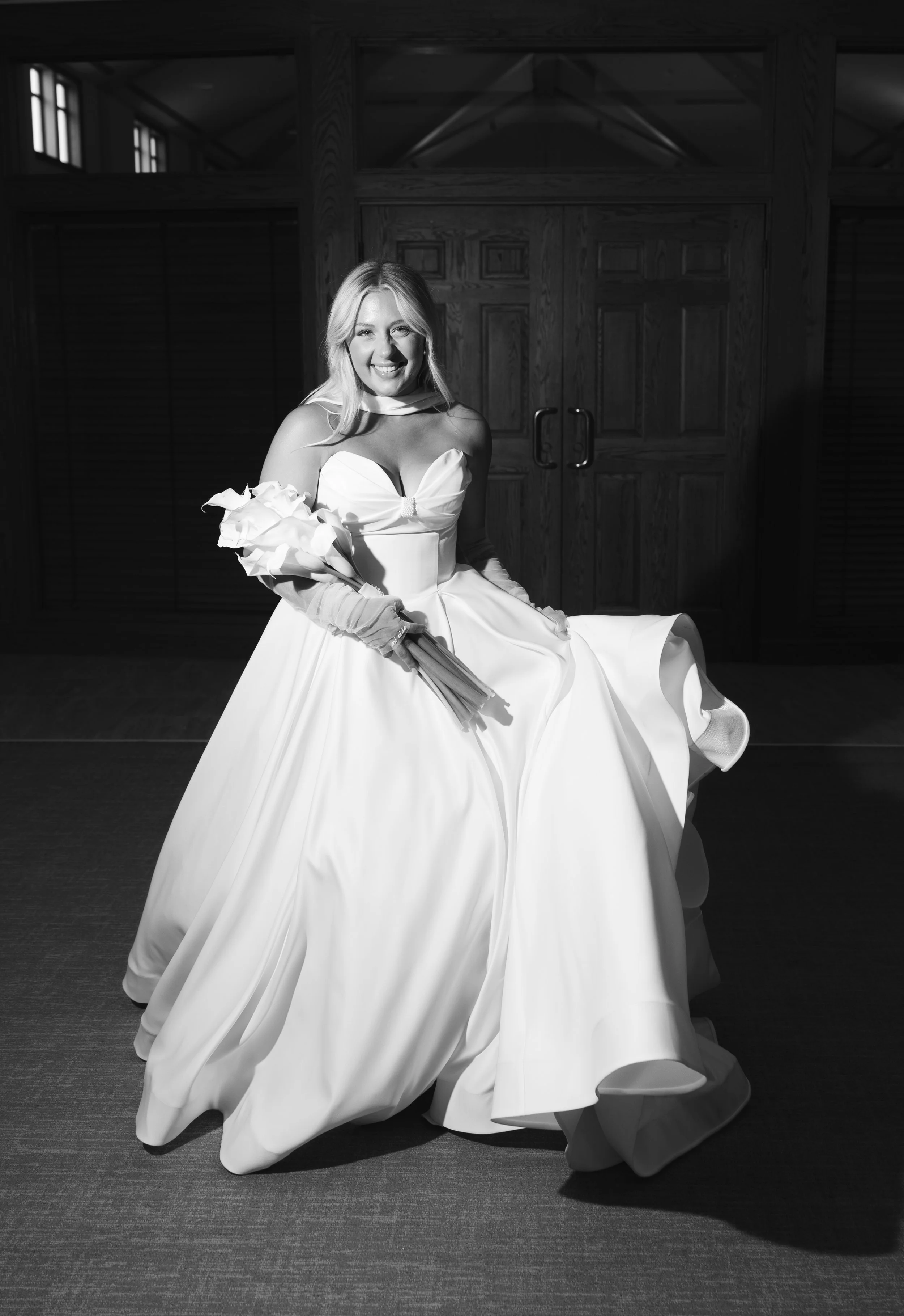 A woman in a wedding dress sitting indoors, smiling, holding a bouquet of flowers.