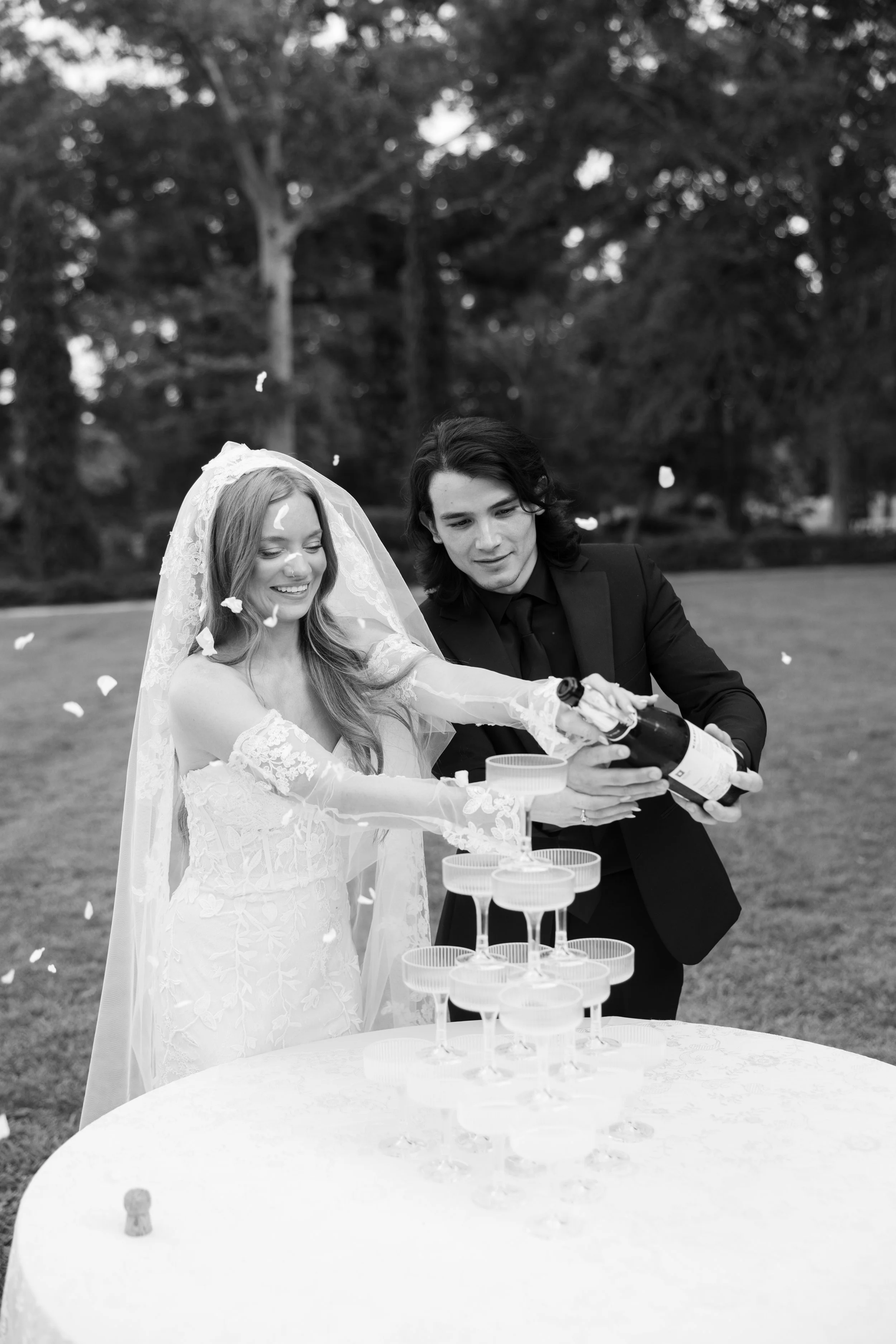 Bride and groom opening champagne over a tower of glasses at an outdoor wedding celebration.