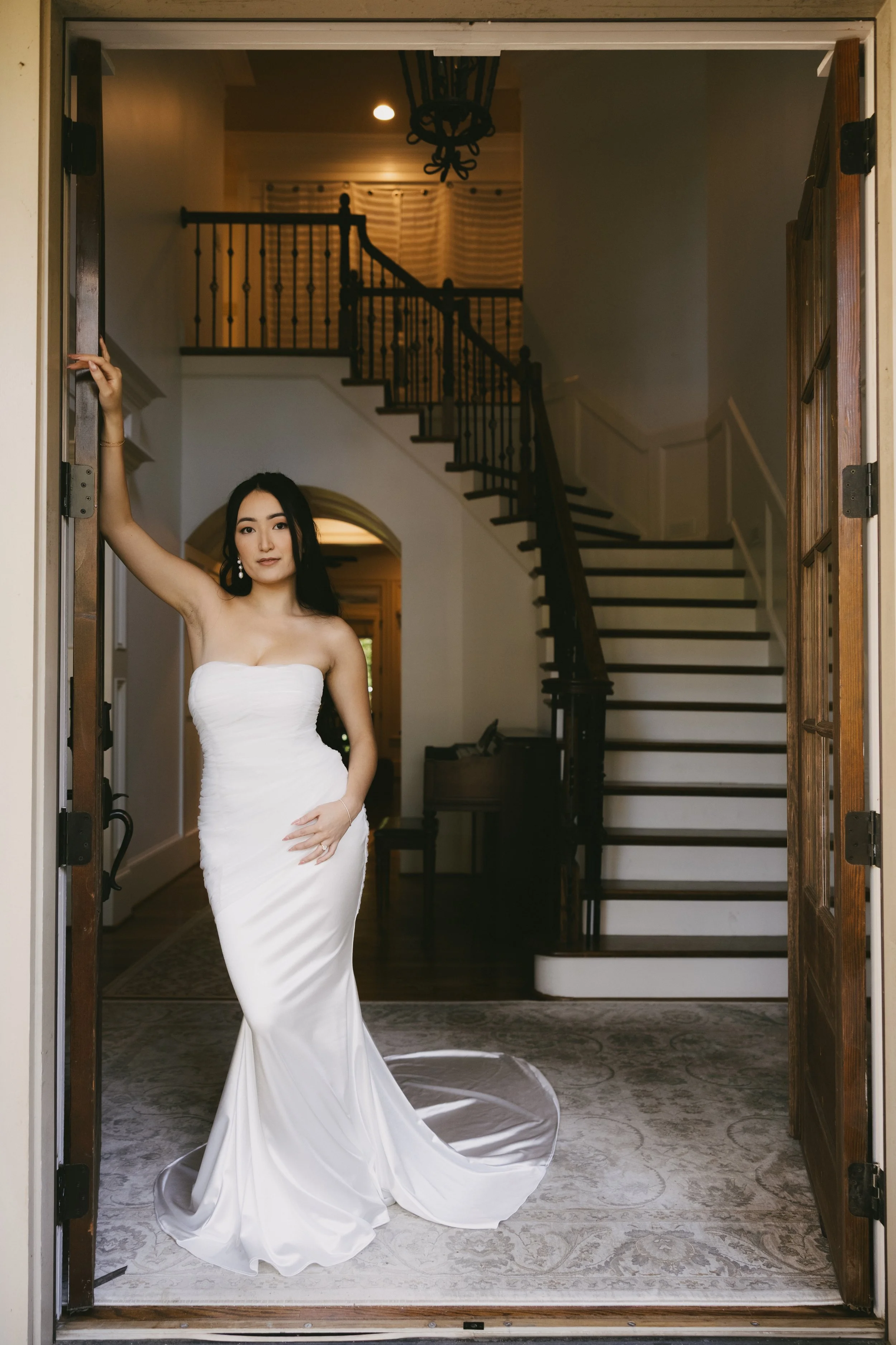 A woman in a white strapless wedding dress standing in a doorway, inside a house with a staircase in the background.