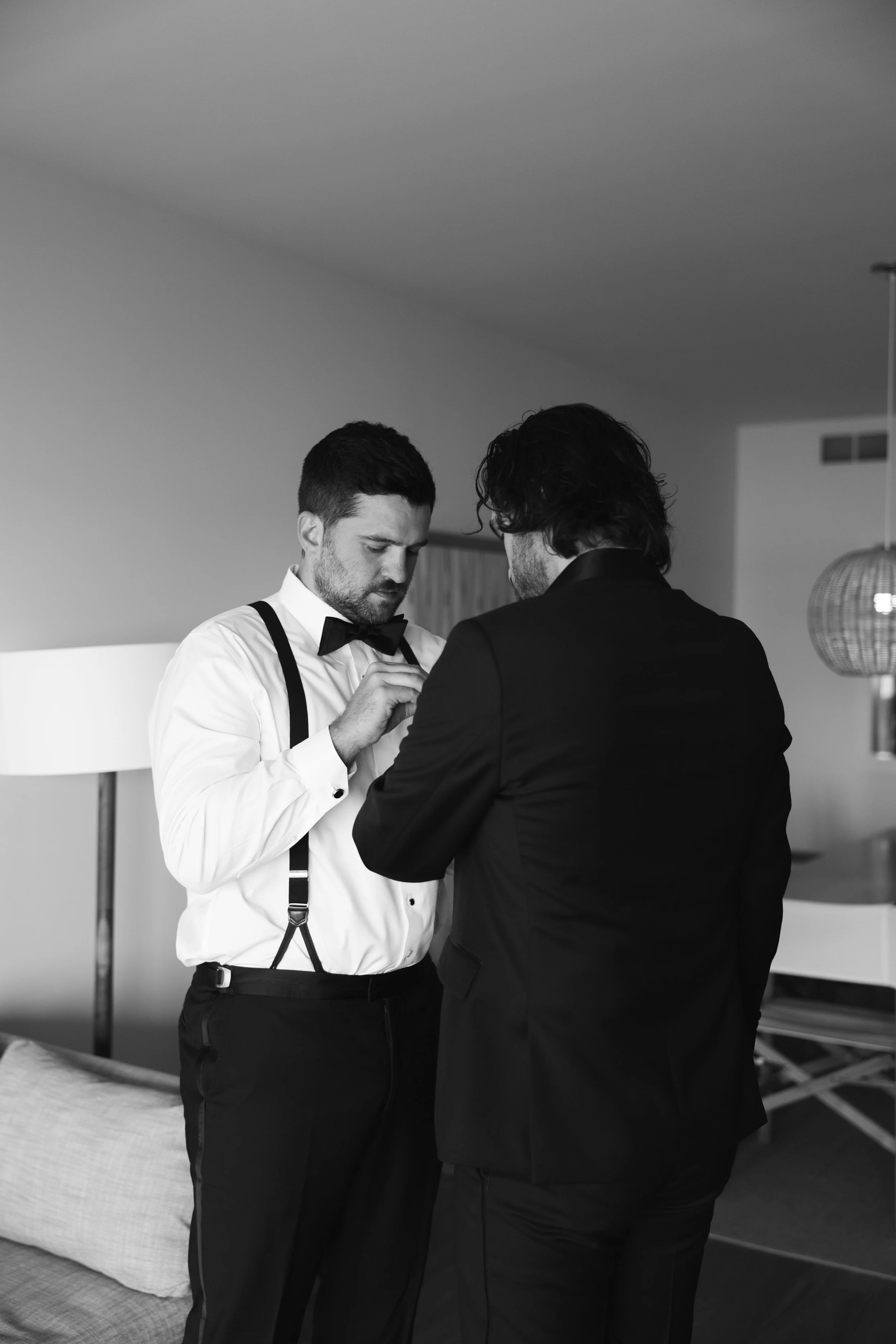 Two men dressed in formalwear standing close together in a room, one adjusting the other's bow tie.