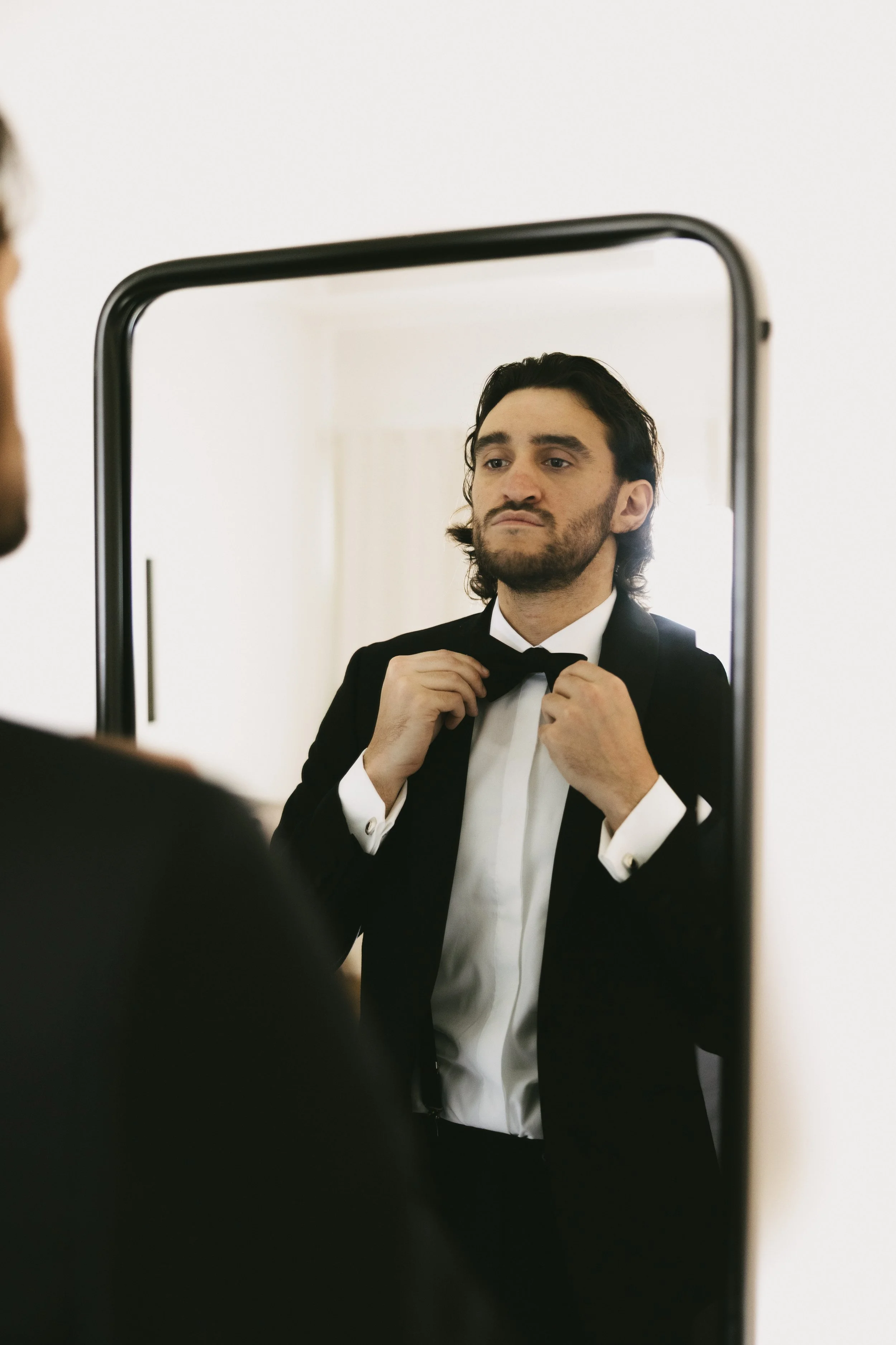 Man in a tuxedo adjusting his bow tie while looking in a mirror.