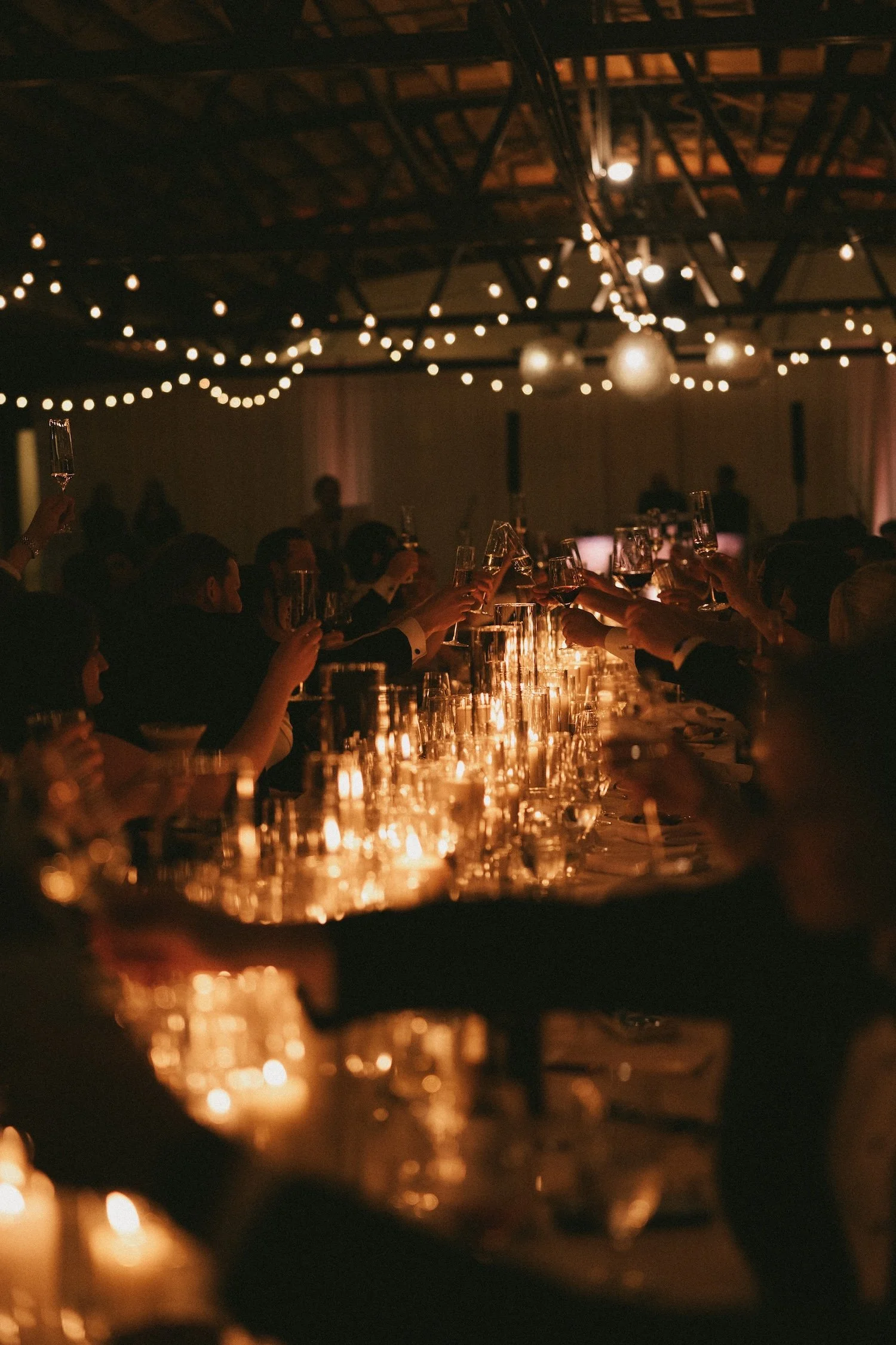 People raising glasses in a toast at a formal dinner party with warm candlelight and string lights overhead.