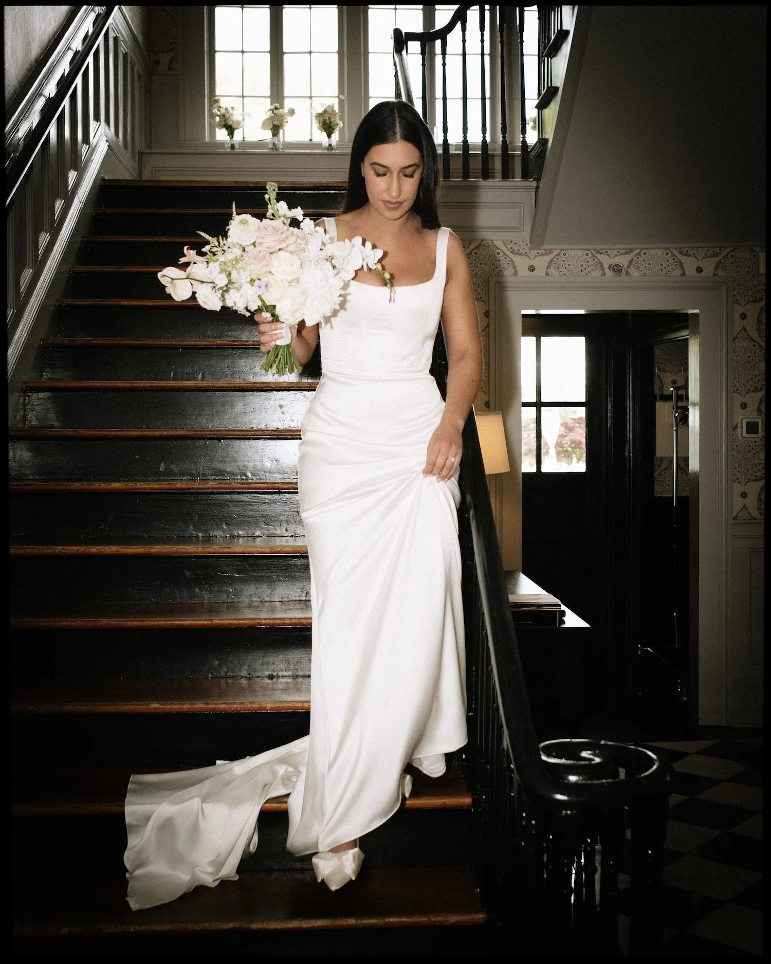 A woman in a white wedding dress holding a bouquet of flowers on a staircase inside a house.