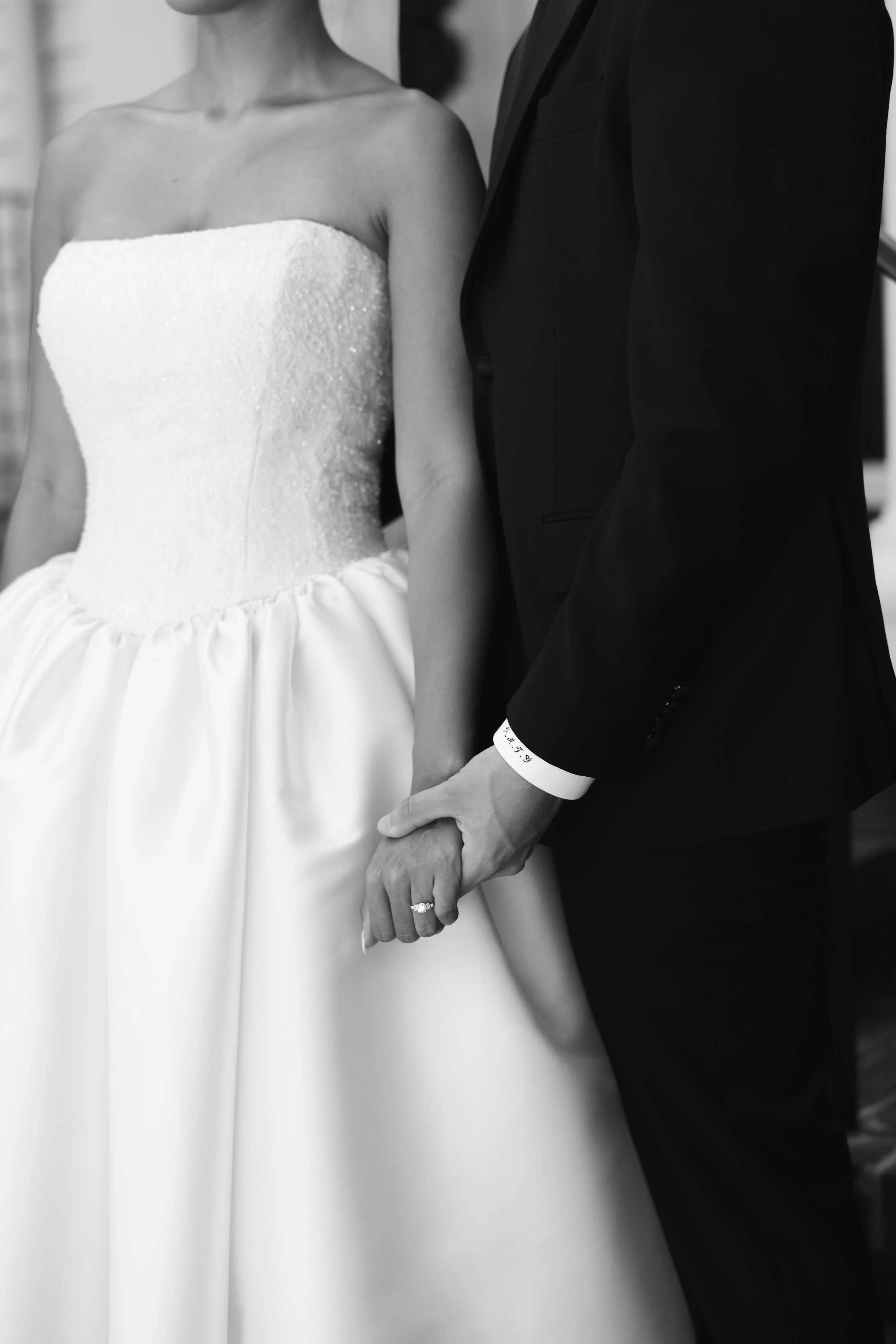 A bride and groom holding hands, with the bride in a strapless wedding gown and the groom in a black suit, focusing on their joined hands and wedding rings.