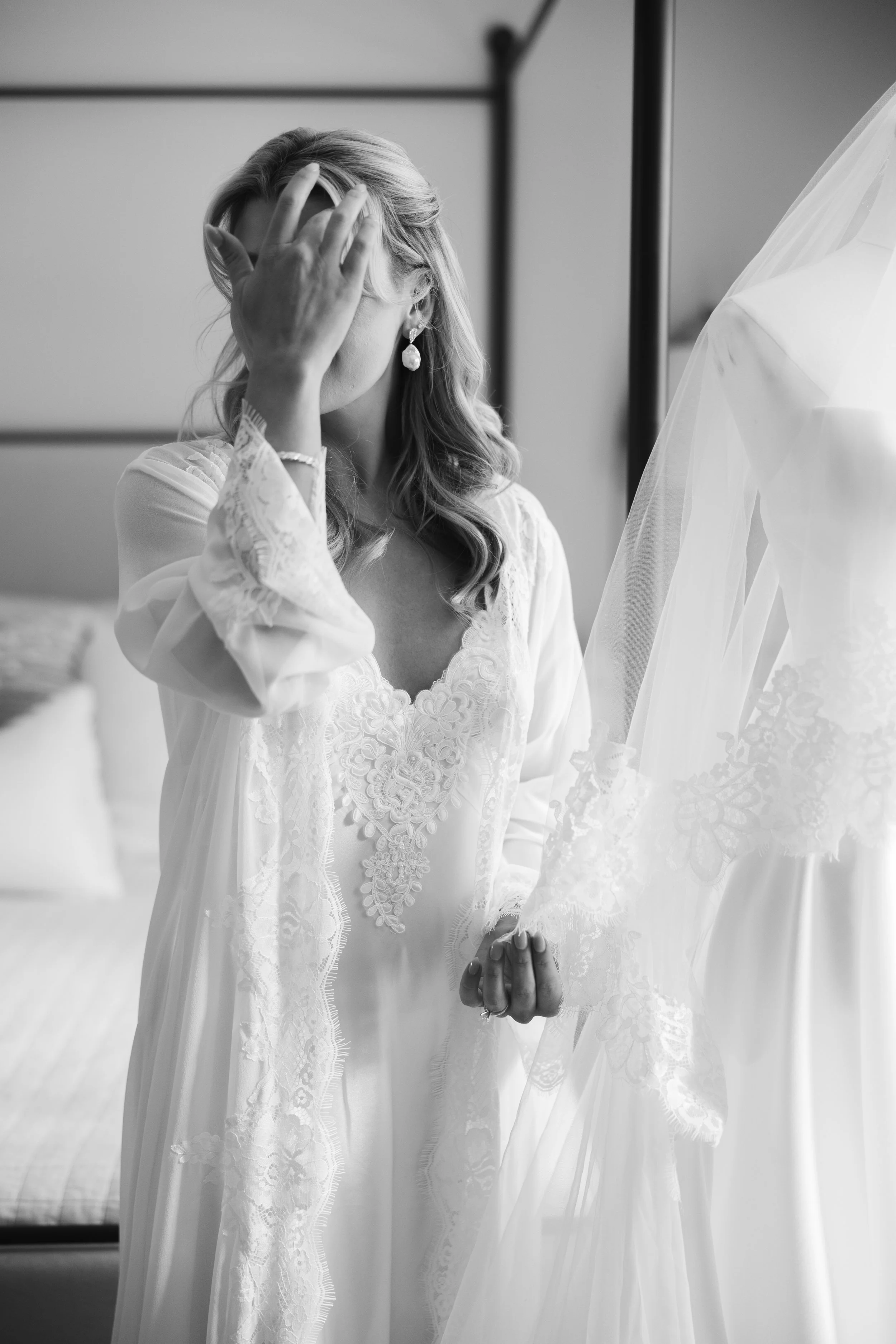 A woman in a lace-trimmed white robe standing in front of a wedding dress on a mannequin, touching her forehead with one hand and holding the dress with the other, in a room with a mirror.