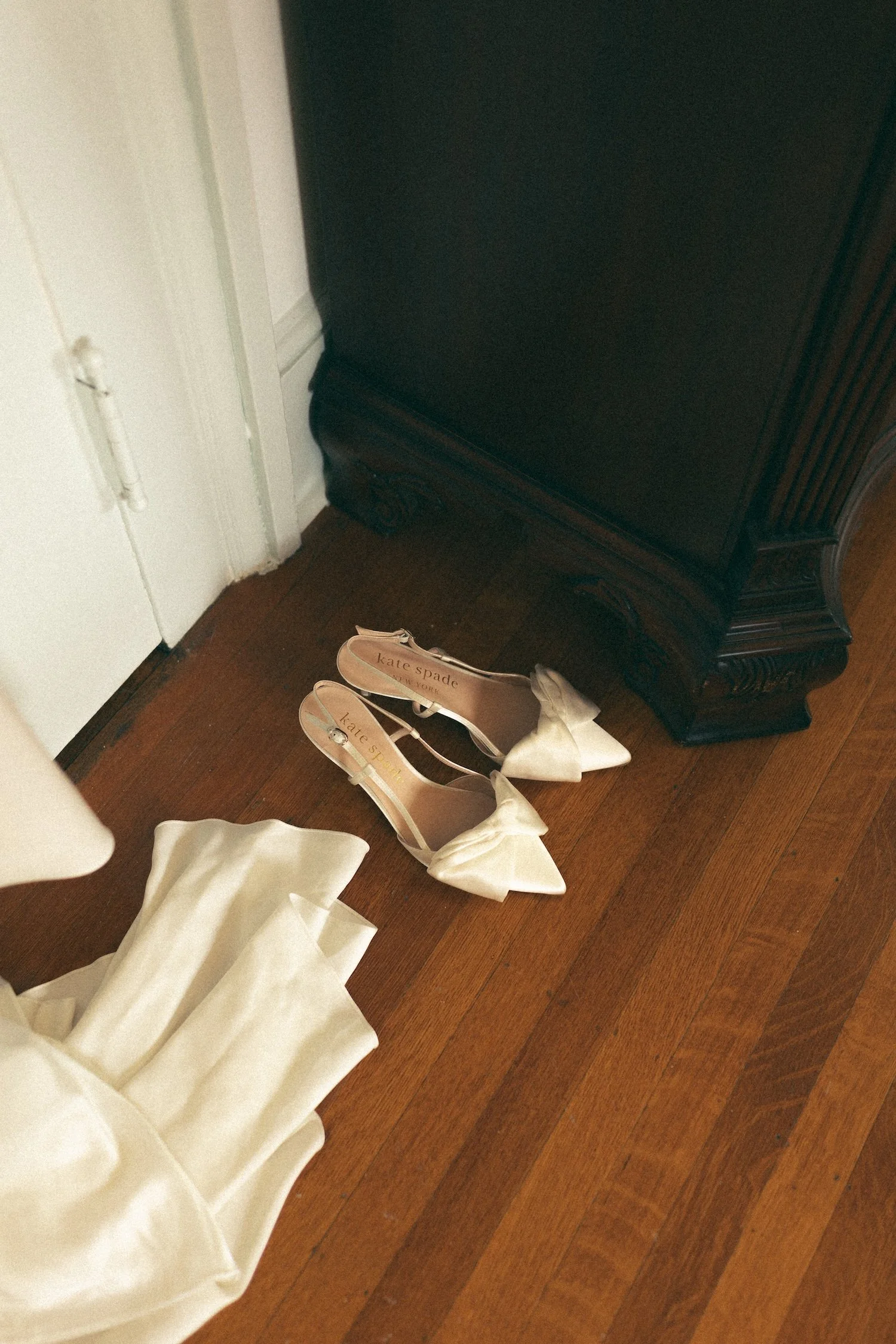Pair of white Kate Spade high-heeled shoes with bows on the toes placed on a hardwood floor.