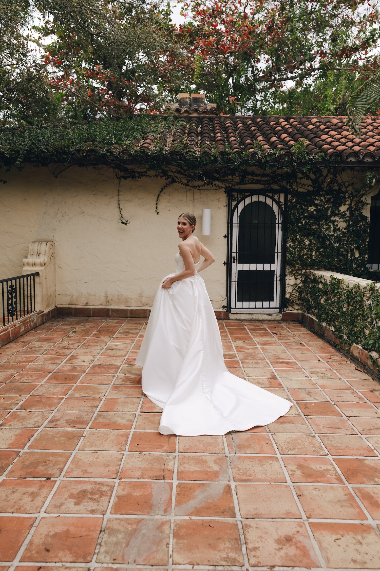 Bride in a white wedding dress standing on terracotta tiled patio, smiling and looking back, in front of a cream wall with vine-covered door and greenery, outdoors.