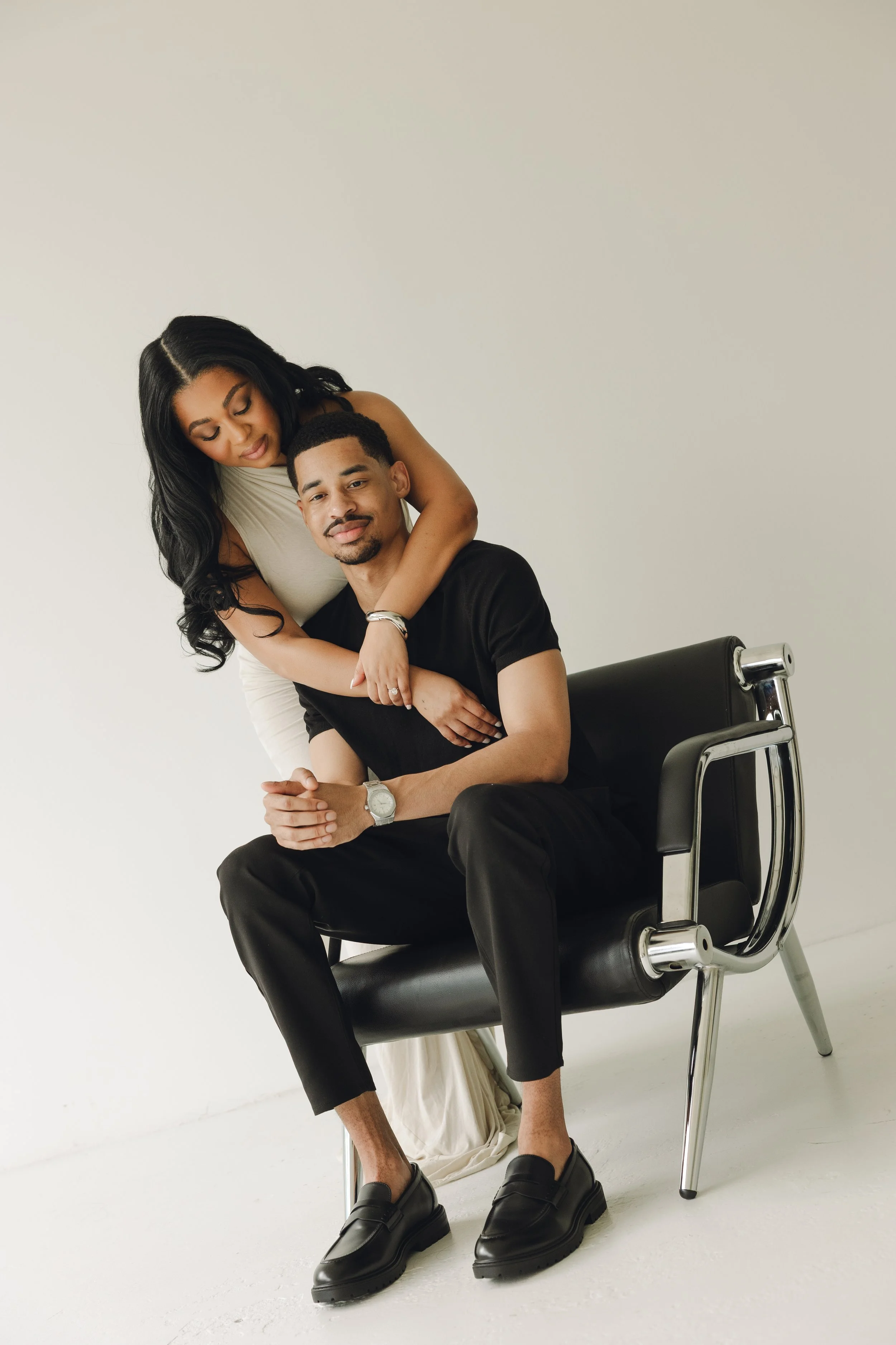 A woman hugging a man sitting on a black chair against a plain white background.