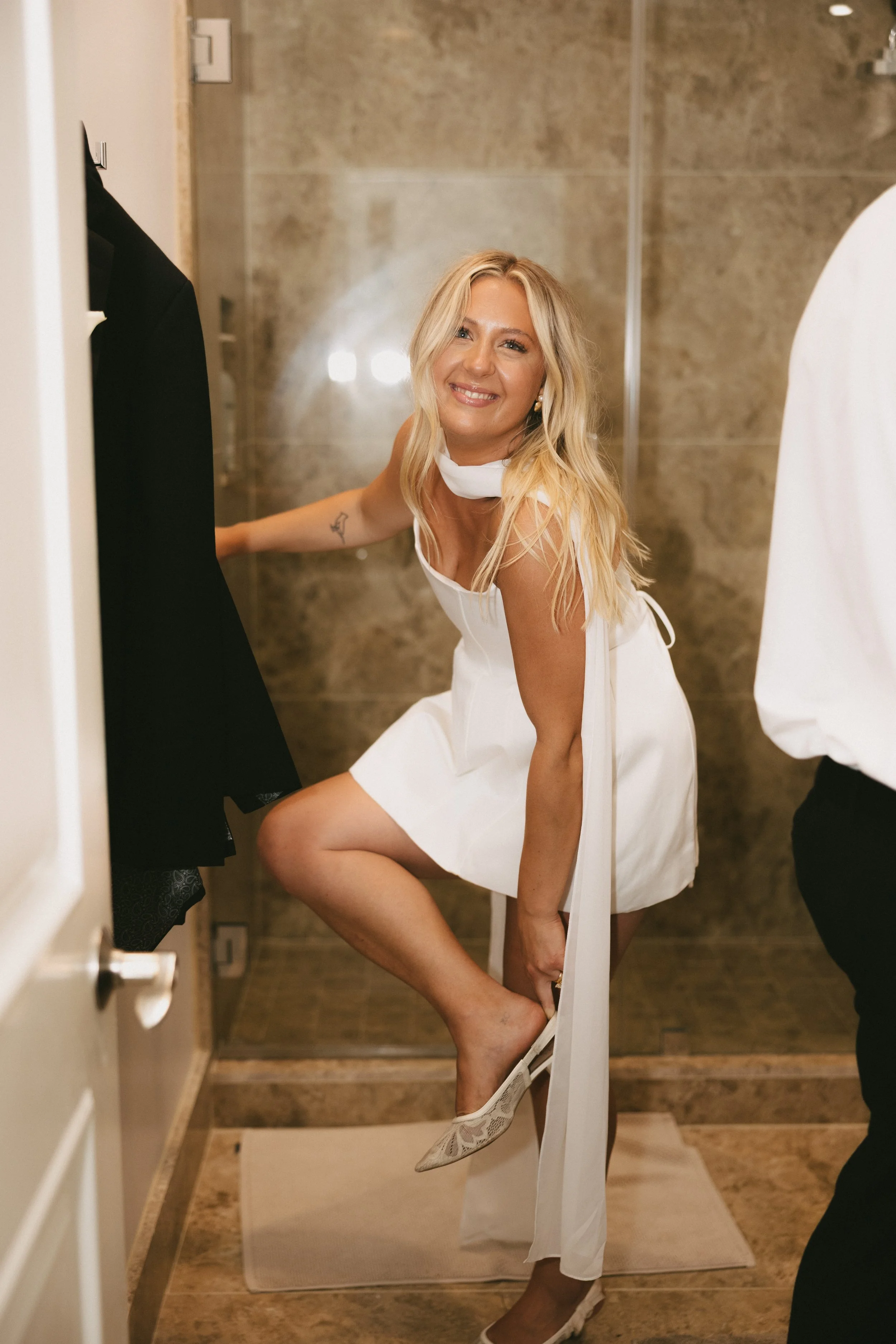A woman in a white dress is squatting and putting on a shoe in a bathroom with beige tiles, smiling at the camera.