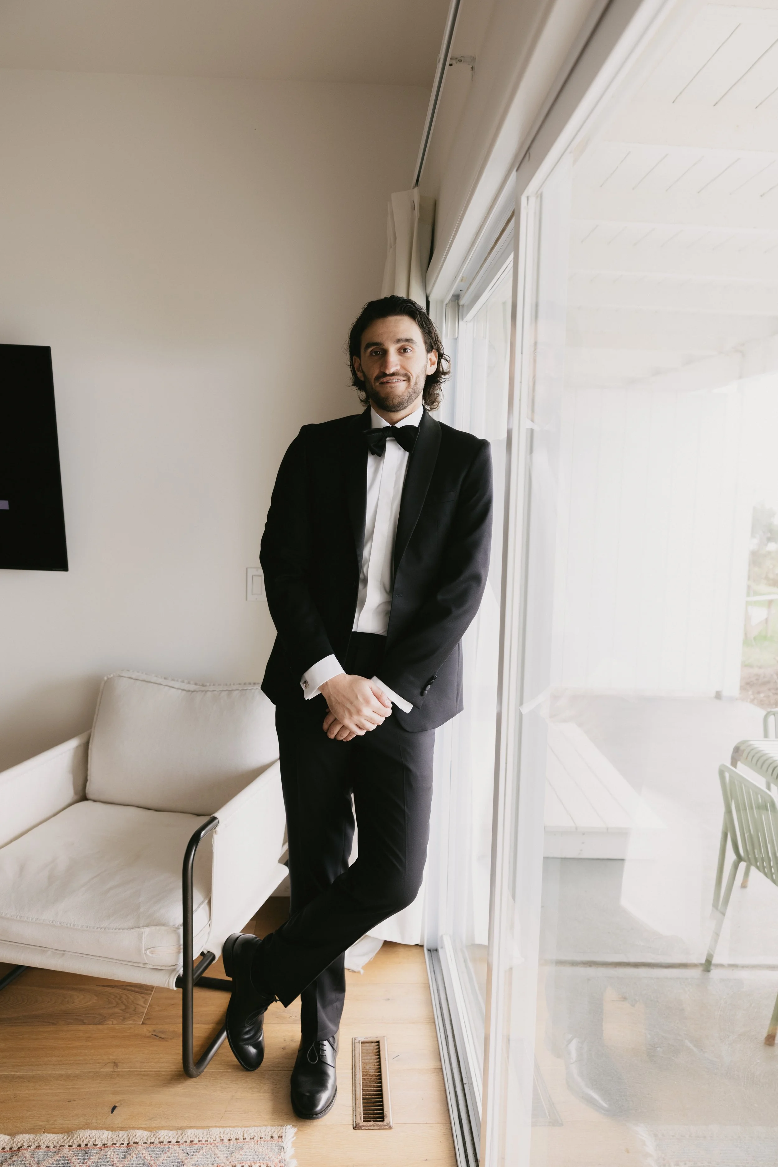 A man in a black tuxedo with a bow tie standing near sliding glass doors, smiling with one foot crossed over the other.