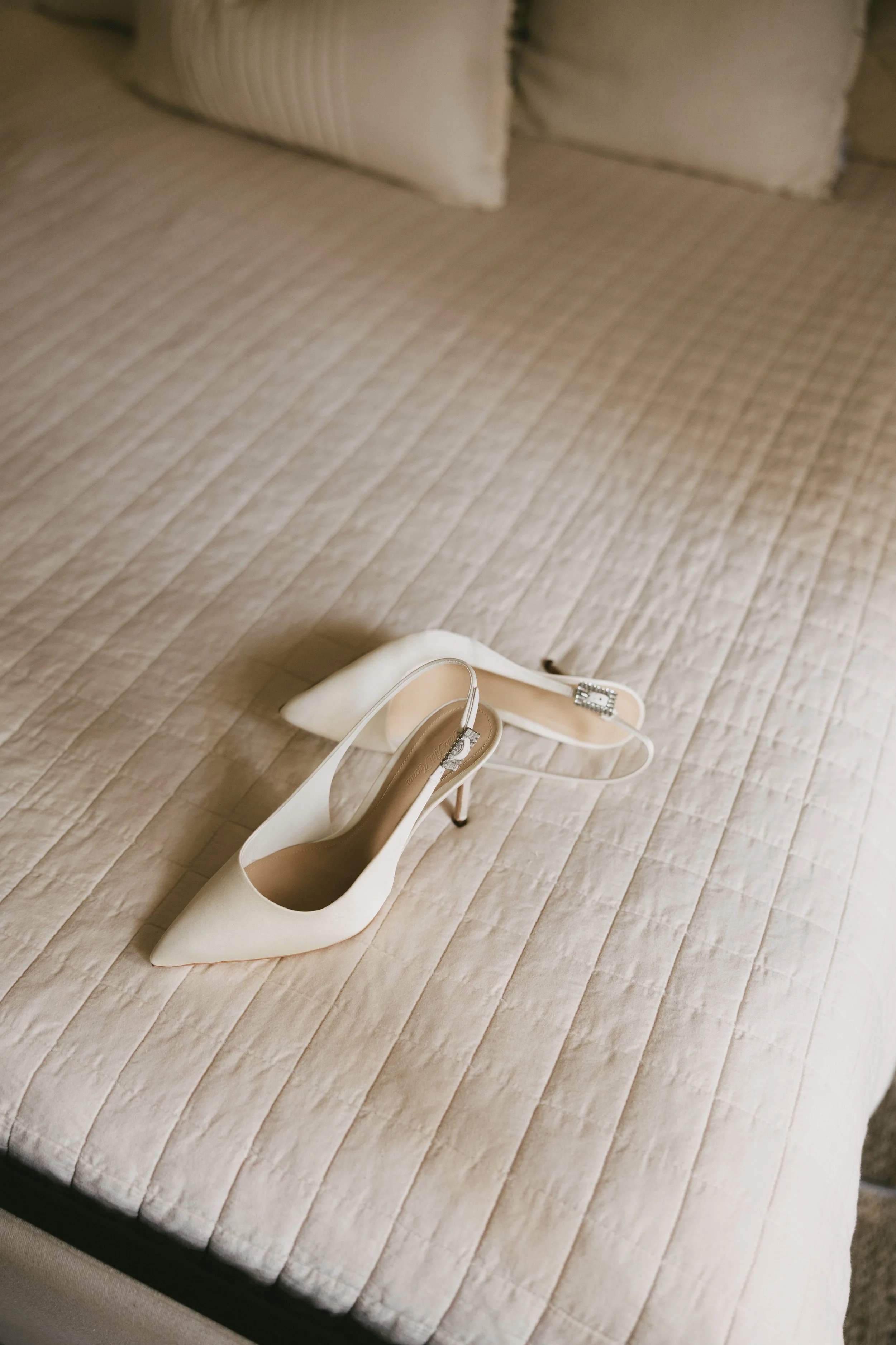 A pair of white high-heeled shoes with ankle straps and decorative buckles resting on a beige quilted bedspread.
