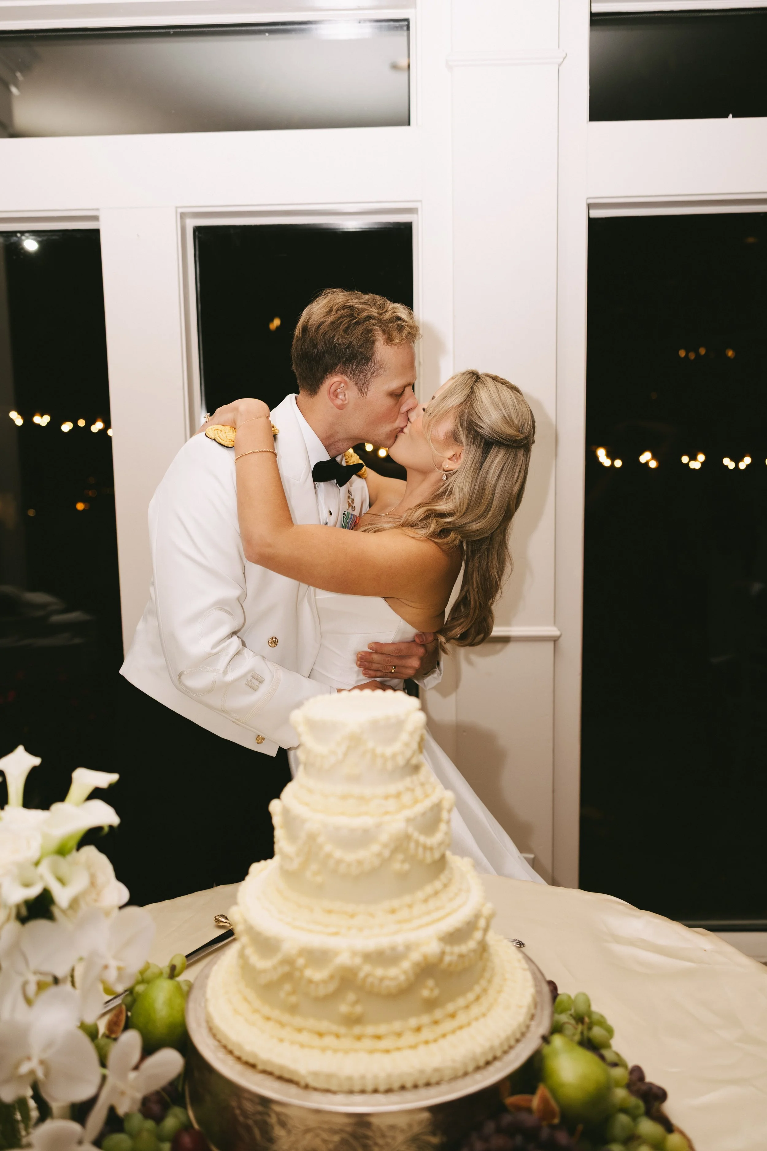 A couple dressed in a wedding gown and military uniform kissing at their wedding reception with a tiered wedding cake in the foreground.