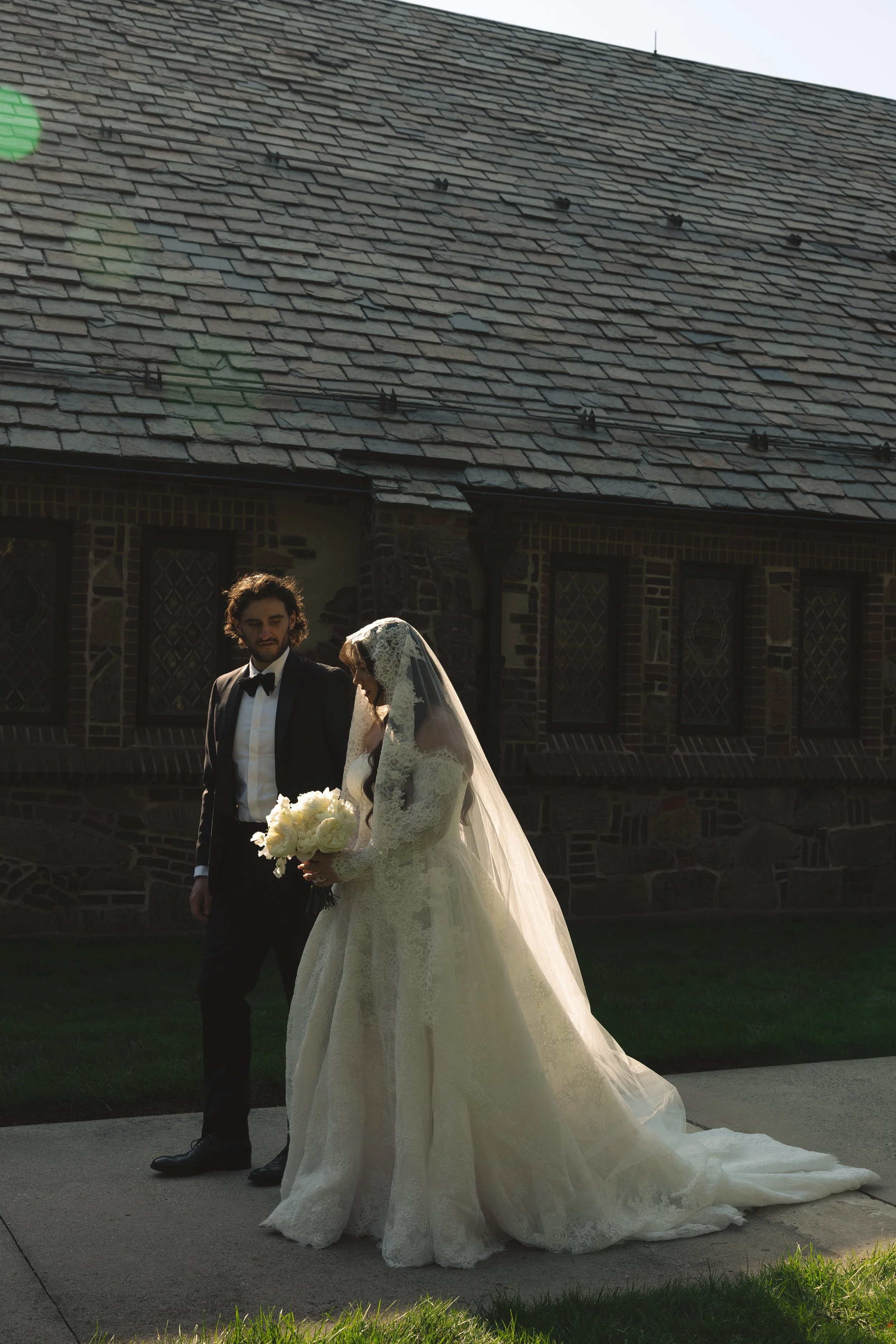 A bride and groom standing outdoors, with the bride holding a bouquet of white flowers, near an old brick building with a slate roof.