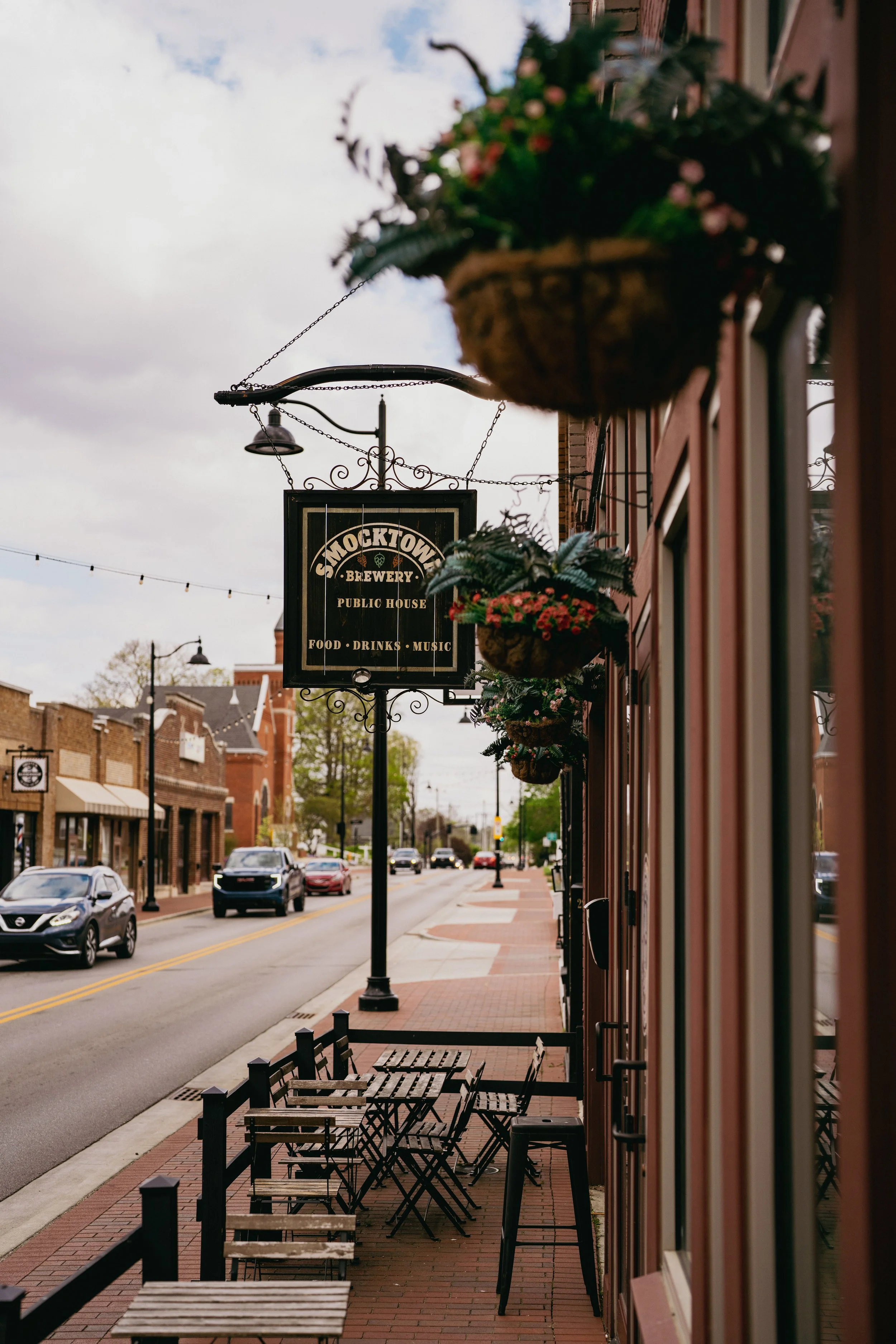 Empty outdoor sidewalk seating area with chairs and tables in front of a restaurant, with hanging flower baskets, along a street with cars and brick buildings.