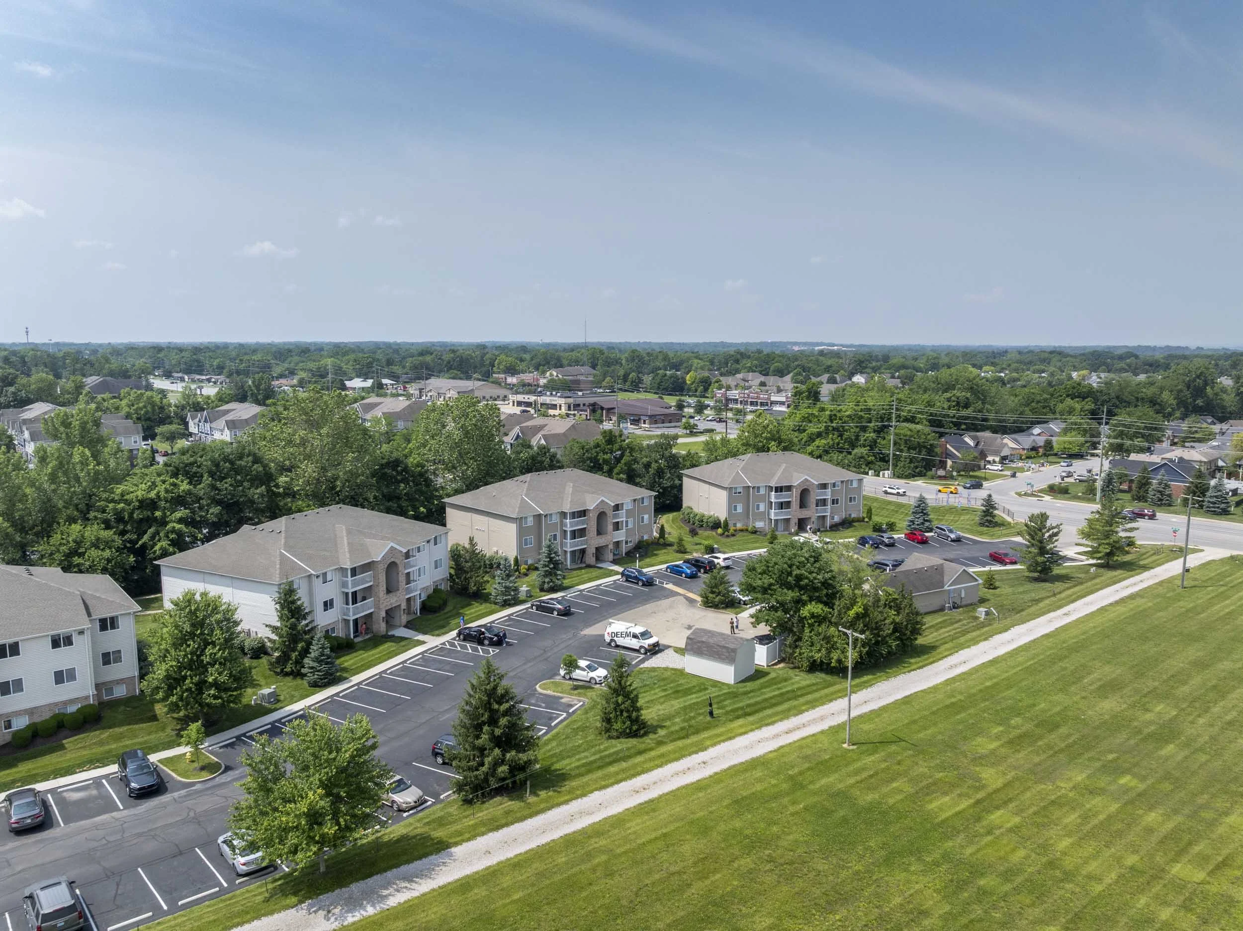 Aerial view of a suburban neighborhood with apartment buildings, a parking lot, trees, and green lawns under a blue sky.
