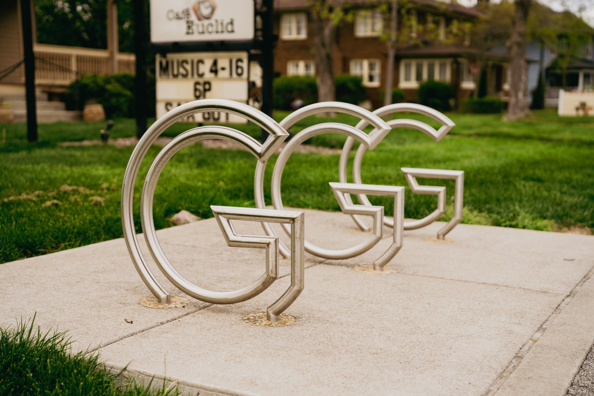 A sidewalk with four metal bike racks in the shape of circles and squares. In the background, there is a sign with the words "Cared Build" and some blurred houses and trees.