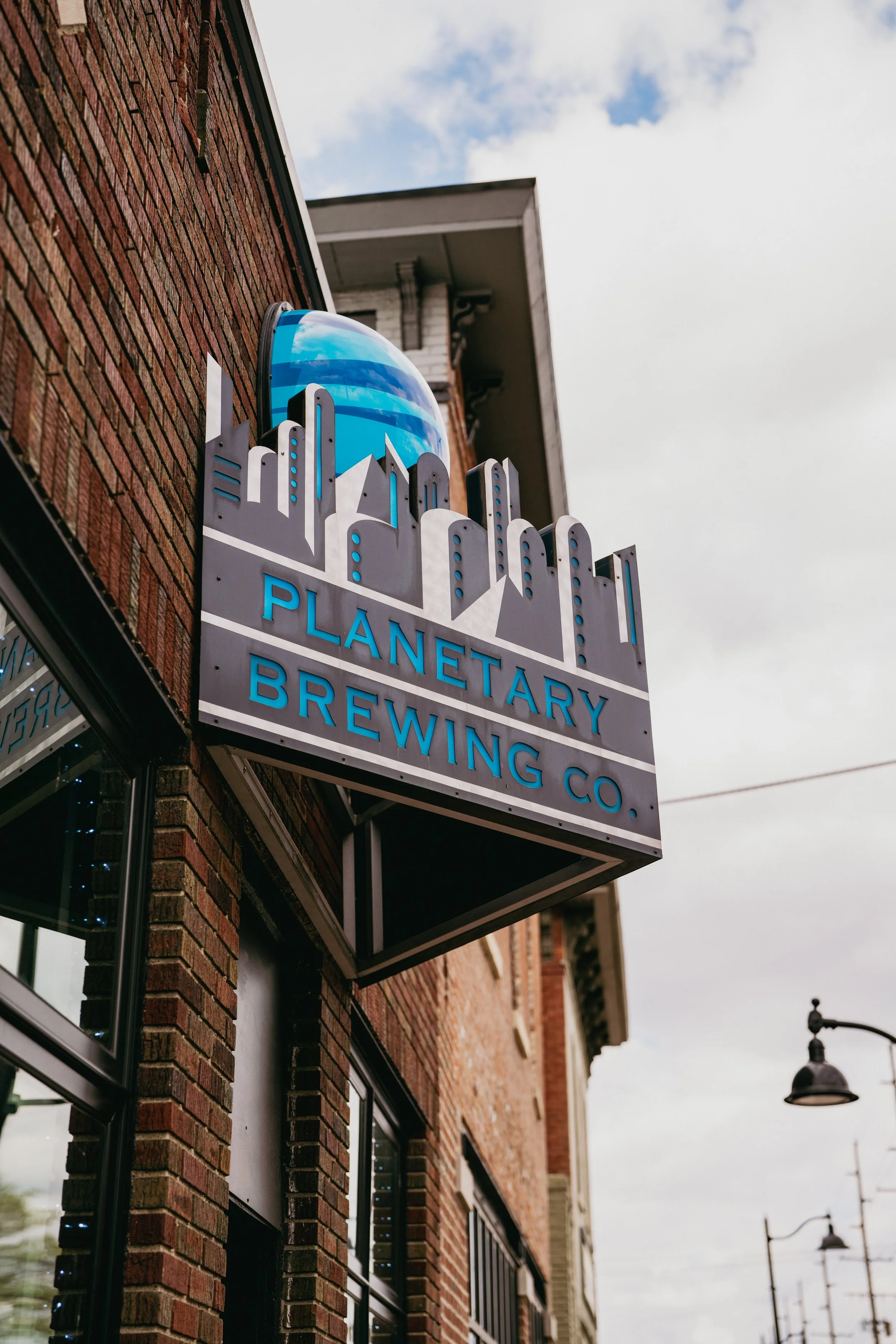 Sign for Planetary Brewing Co. mounted on brick building exterior, includes a blue globe and city skyline graphic, with a streetlamp and cloudy sky in background.