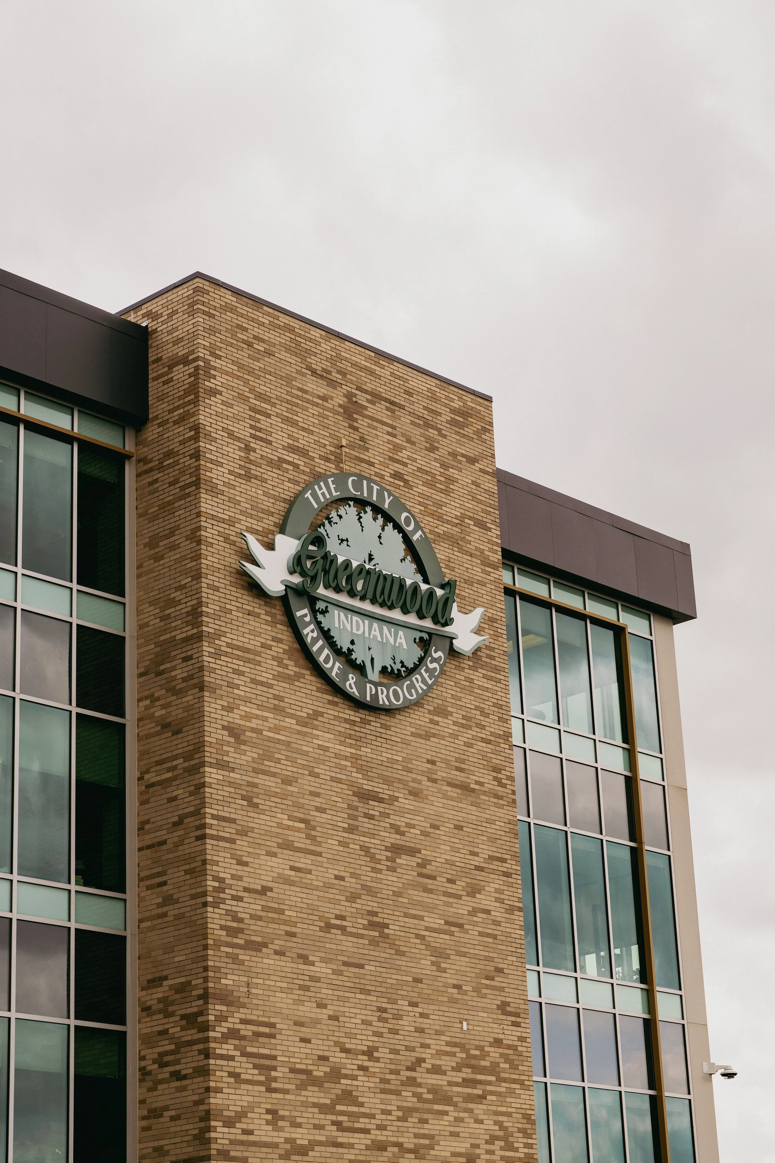 A building with a brick facade and large glass windows, featuring a circular sign that reads 'The City of Greenwood Indiana Pride & Progress' with trees in the background on the sign.