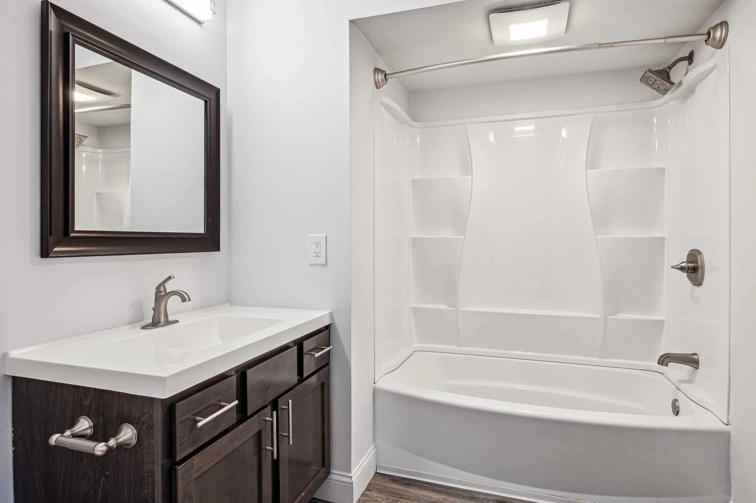 A modern bathroom with a dark wooden vanity, white countertop, mirror, and a shower/tub combo with a showerhead and a curved shower curtain rod.