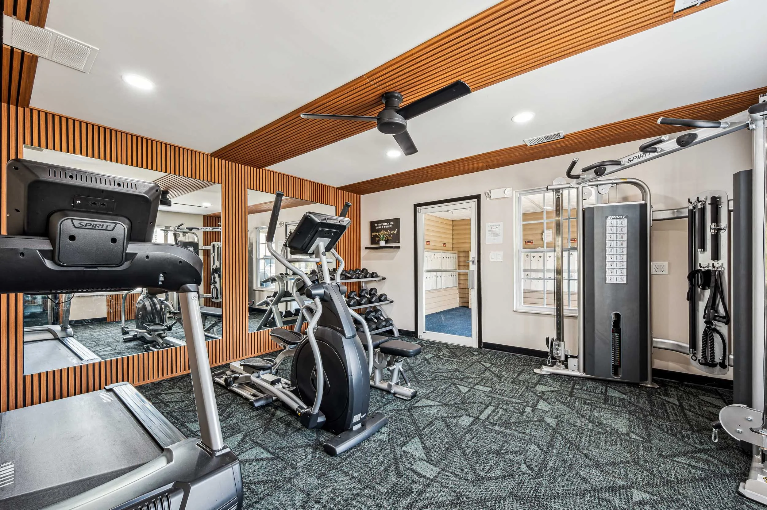 Indoor gym with cardio machines, free weights, and workout equipment, featuring wooden wall accents and a ceiling fan.