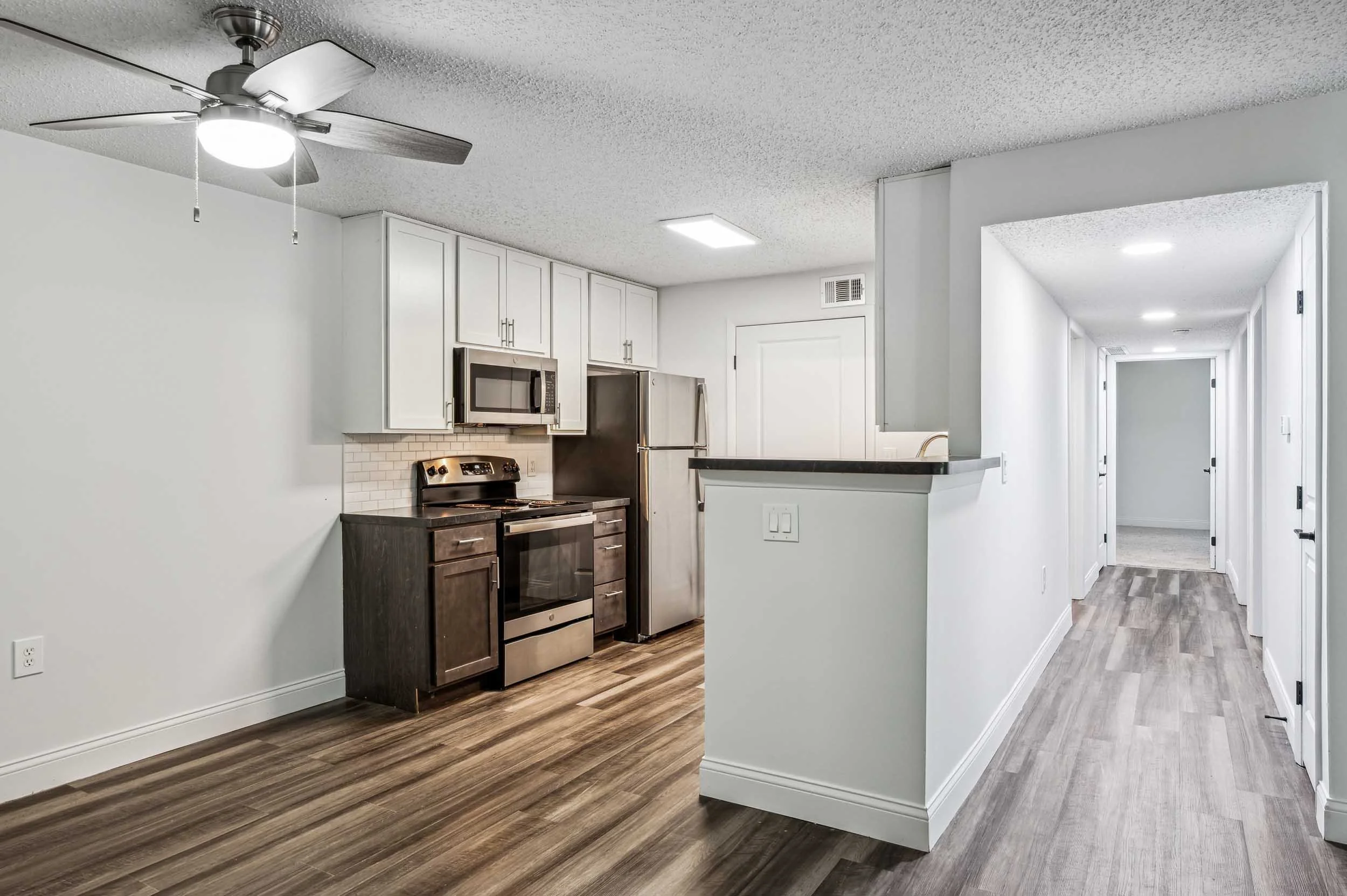 Interior view of a modern kitchen with white cabinets, stainless steel appliances, wooden flooring, and a hallway leading to other rooms.