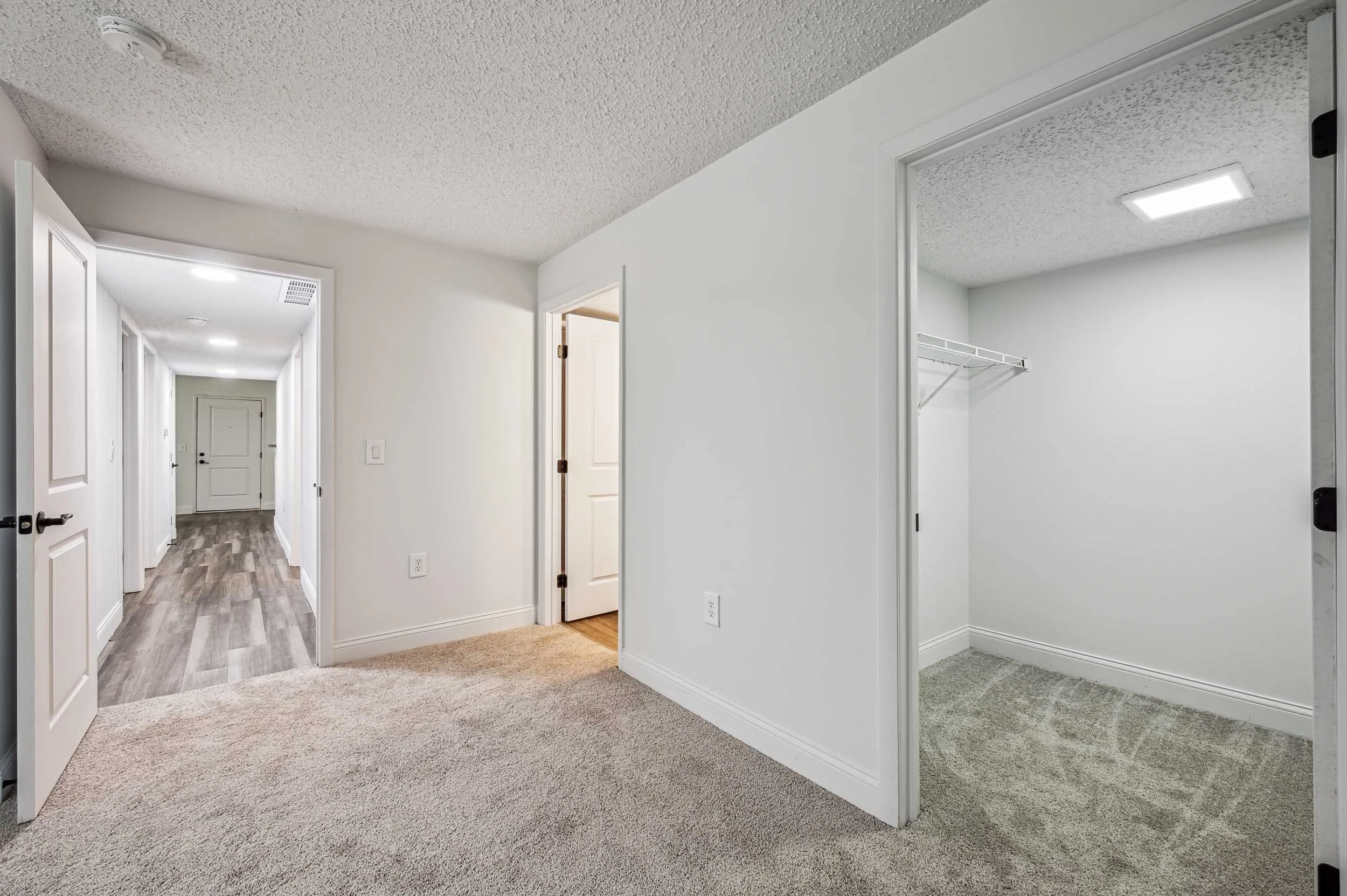 Empty interior space with white walls, carpeted and hardwood flooring, open closet, and open doorway leading to a hallway.