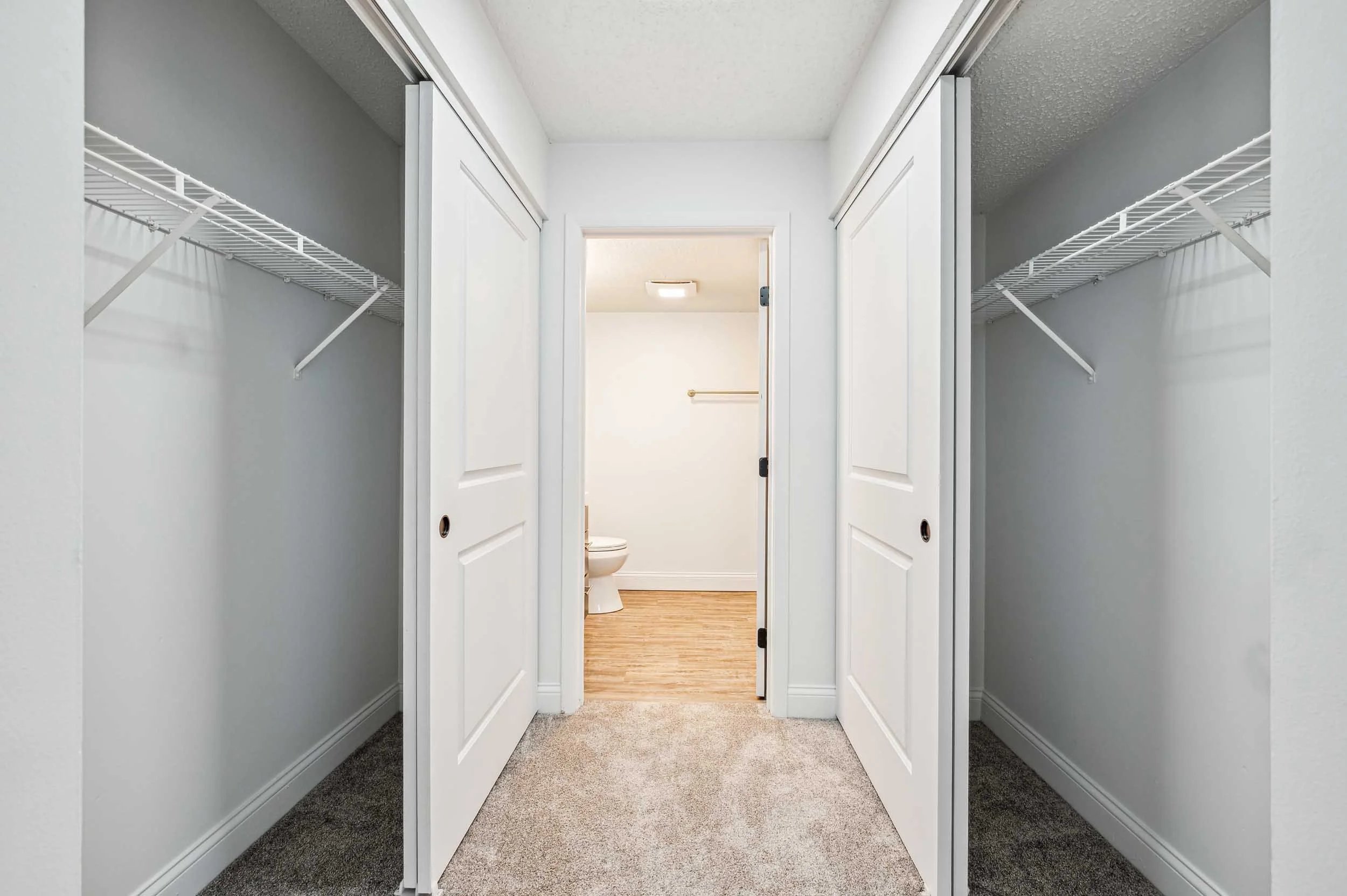 Empty walk-in closet with white double doors open, revealing a small bathroom with a toilet and a towel bar. Closet has gray walls, wire shelves, and carpets on the floor.