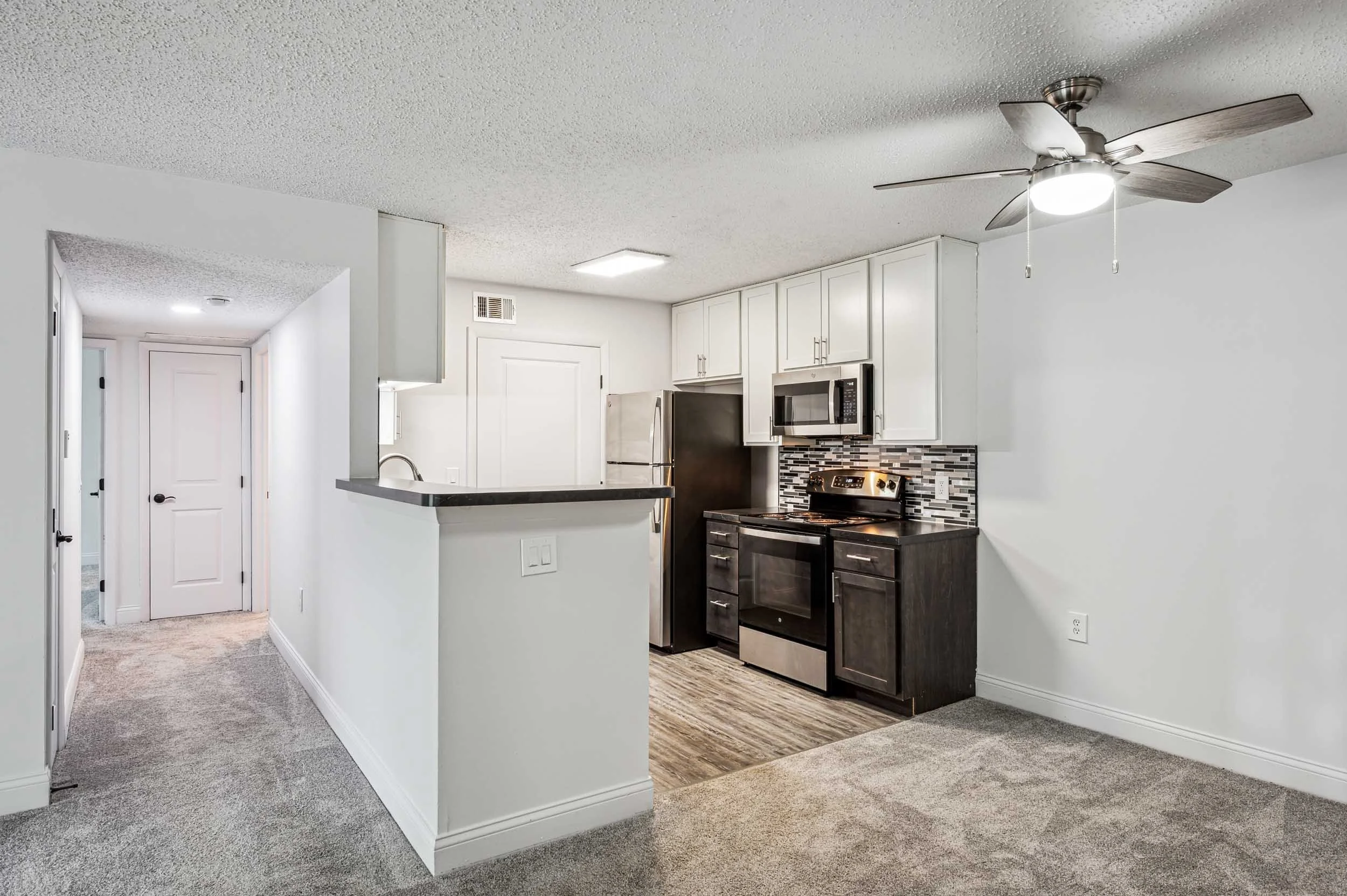 Interior view of a modern kitchen with white cabinets, black stove, microwave, stainless steel refrigerator, and a tiled backsplash, adjacent to a carpeted hallway with white walls and doors.