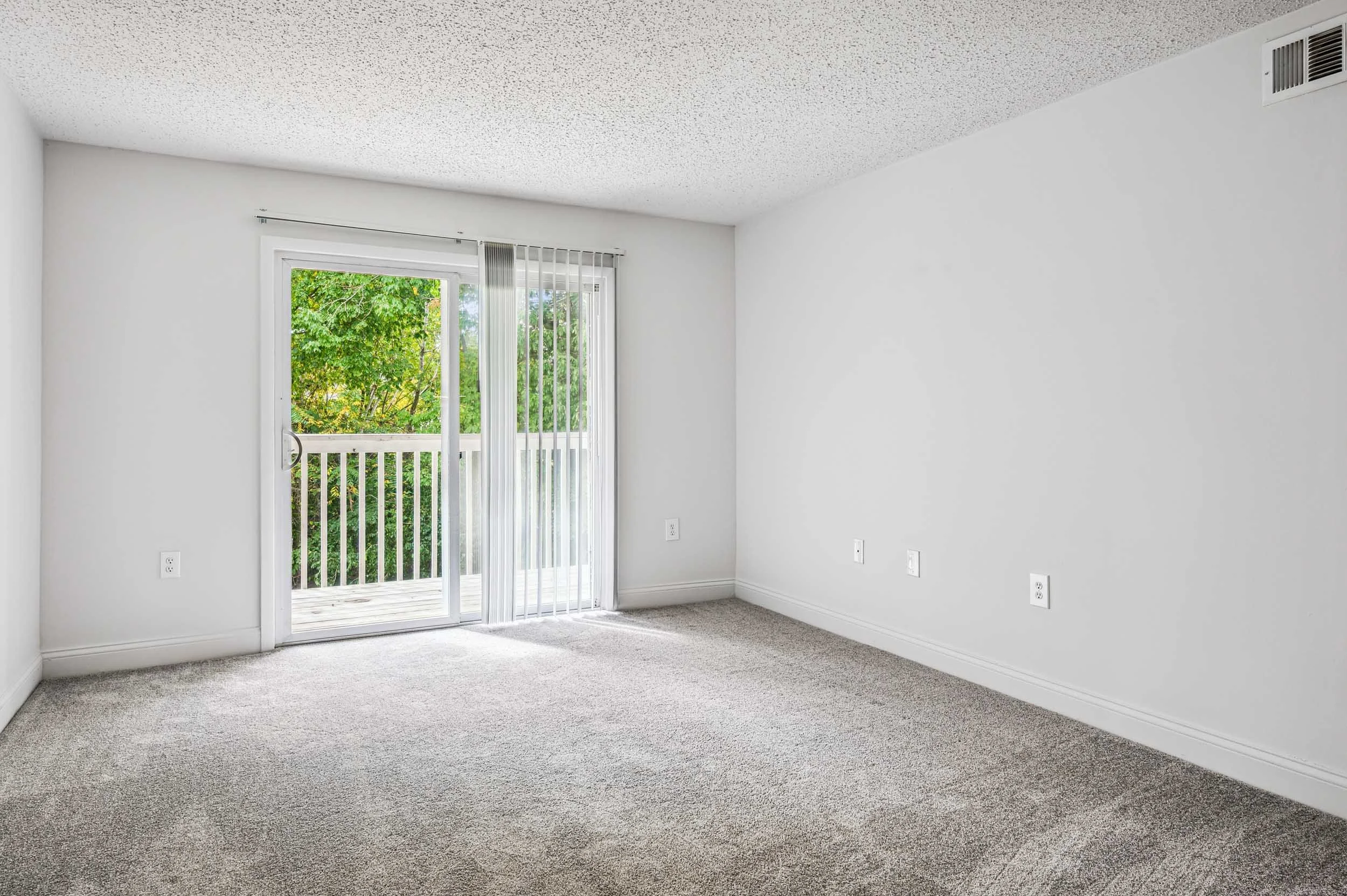 Empty room with beige carpet, white walls, a sliding glass door leading to a balcony, green trees outside, vertical blinds, ceiling vent, multiple electrical outlets.