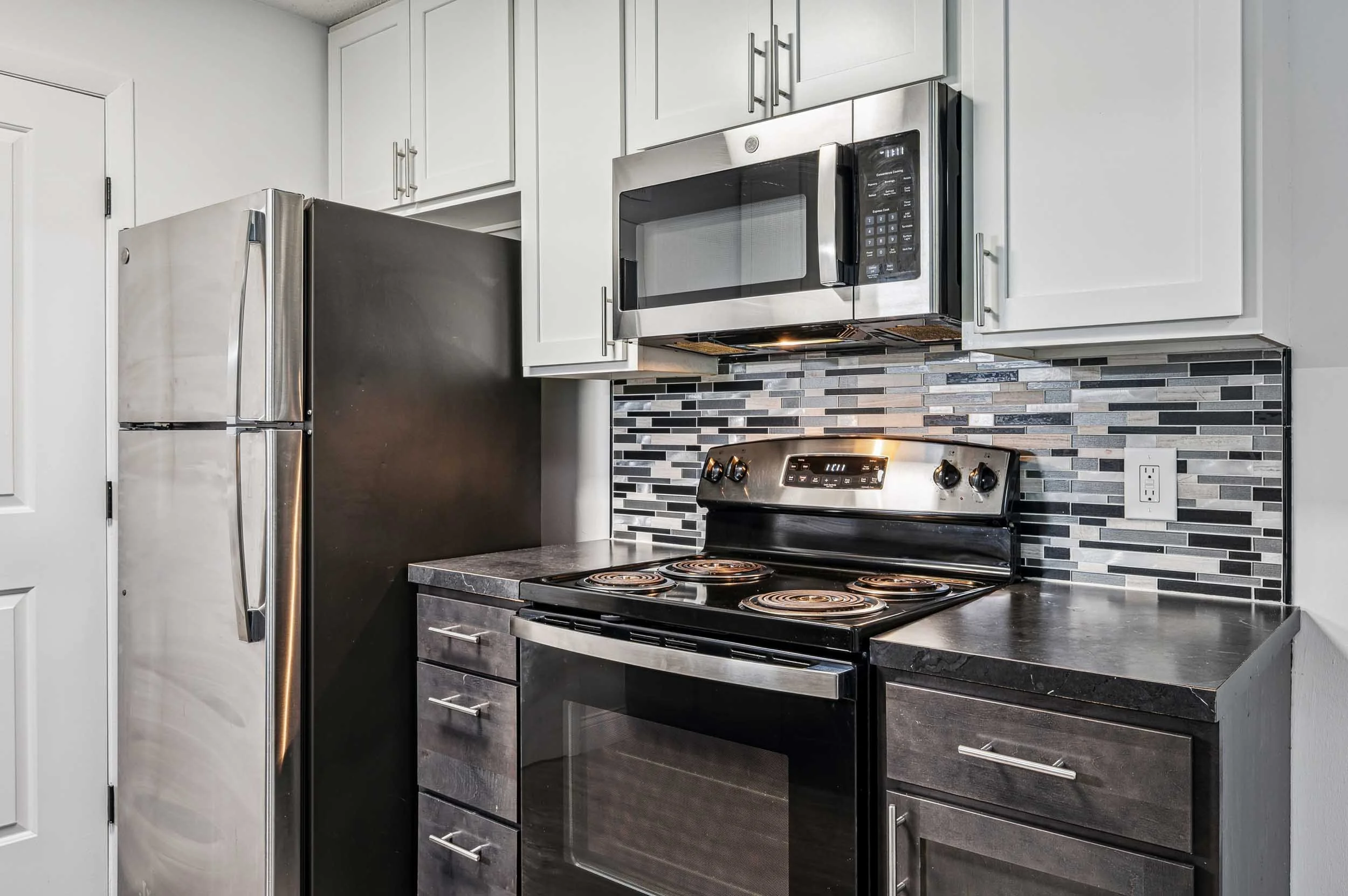 Modern kitchen with stainless steel refrigerator, microwave, and electric stove; black and gray tiled backsplash; white upper cabinets with silver handles; gray granite countertop.