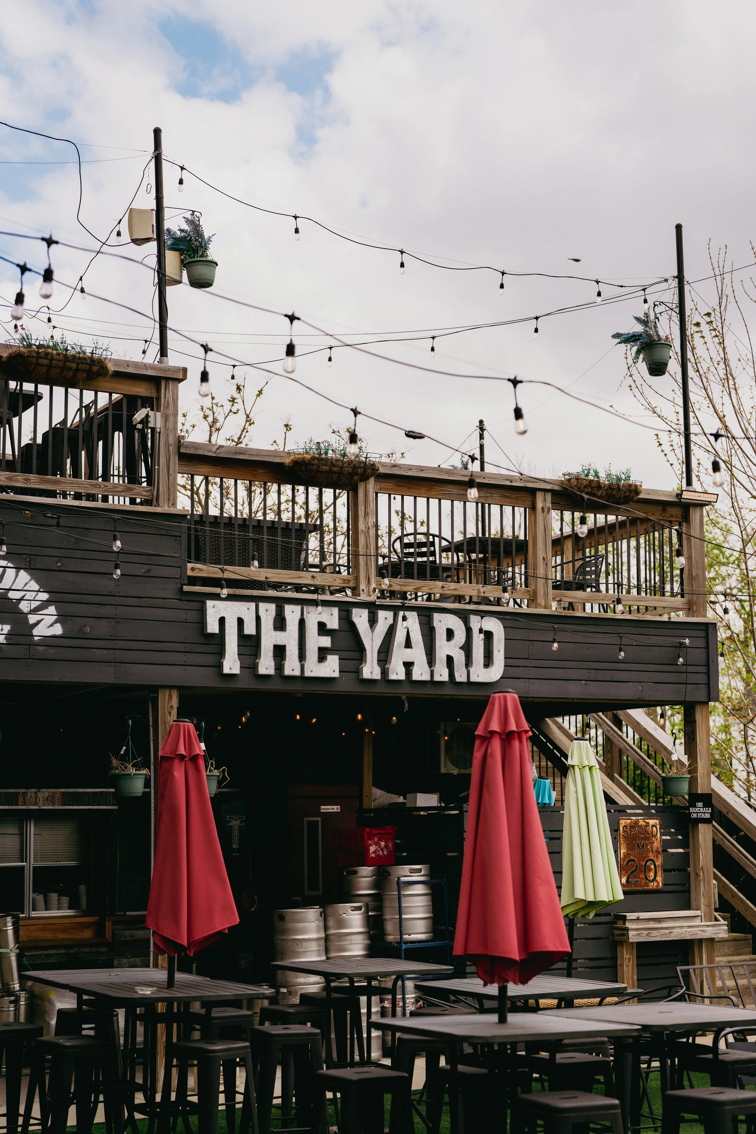Outdoor patio area with string lights, potted plants, red umbrellas, tables, and a sign that reads 'THE YARD'