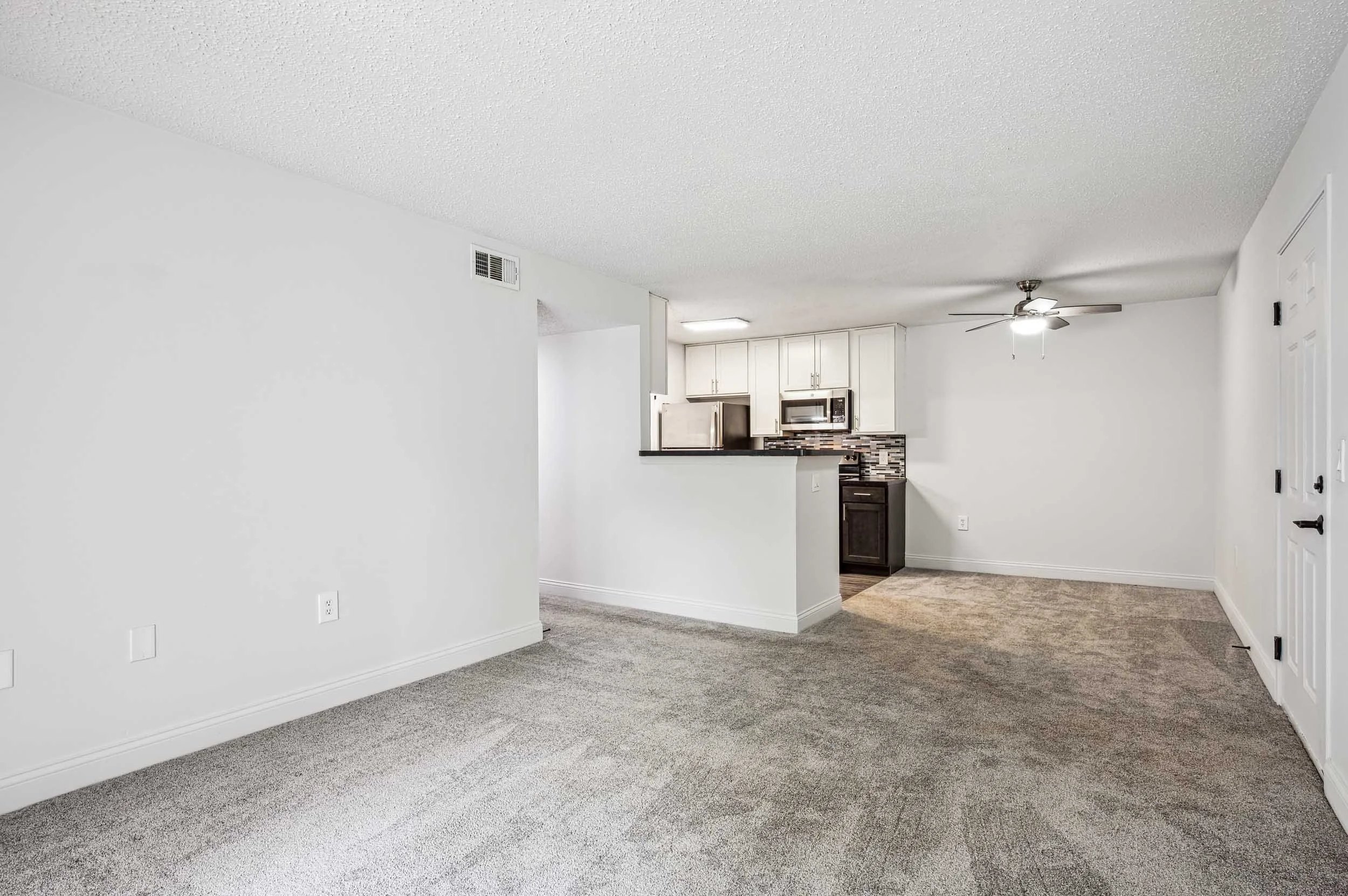 Empty living room with beige carpet, white walls, and a view into a kitchen with white cabinets and a dishwasher.