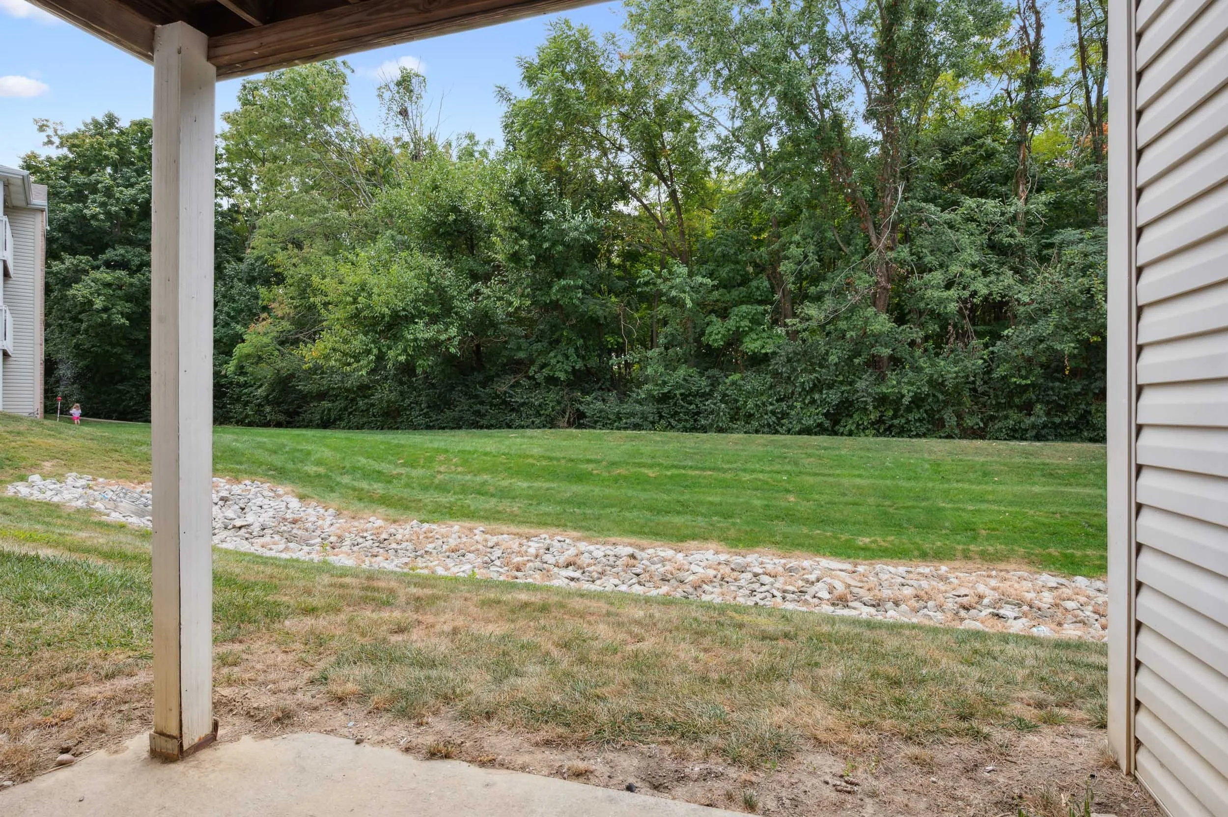 Looking out from a covered patio or porch at a backyard with a grassy lawn, a rock garden, a wooded area with tall green trees, and neighboring houses on the left with siding and balconies.