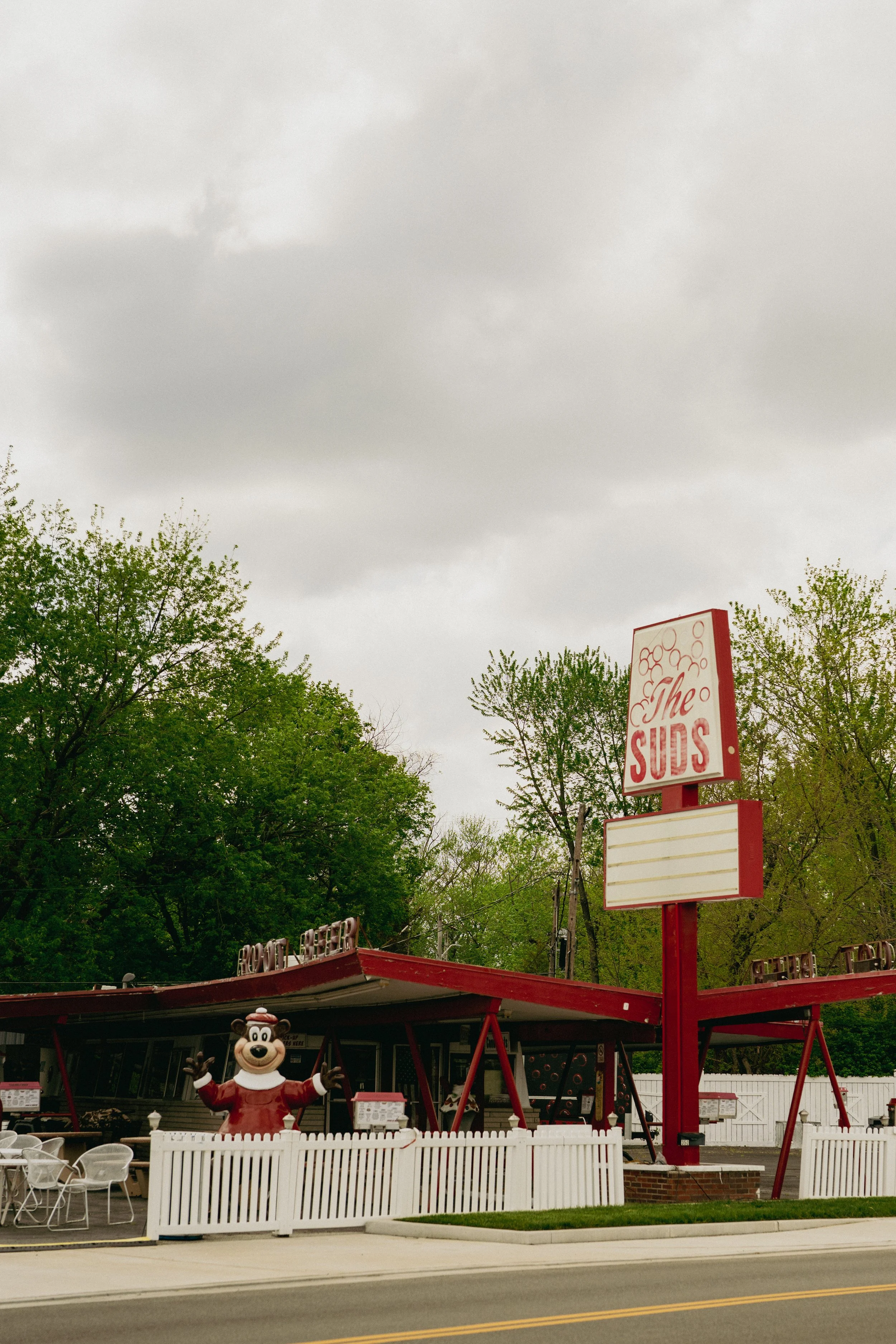 An outdoor restaurant with a red and white sign reading 'The Suds', a mascot figure of a bear in a red shirt and white apron, a white picket fence, tables and chairs, and trees with green leaves under a cloudy sky.