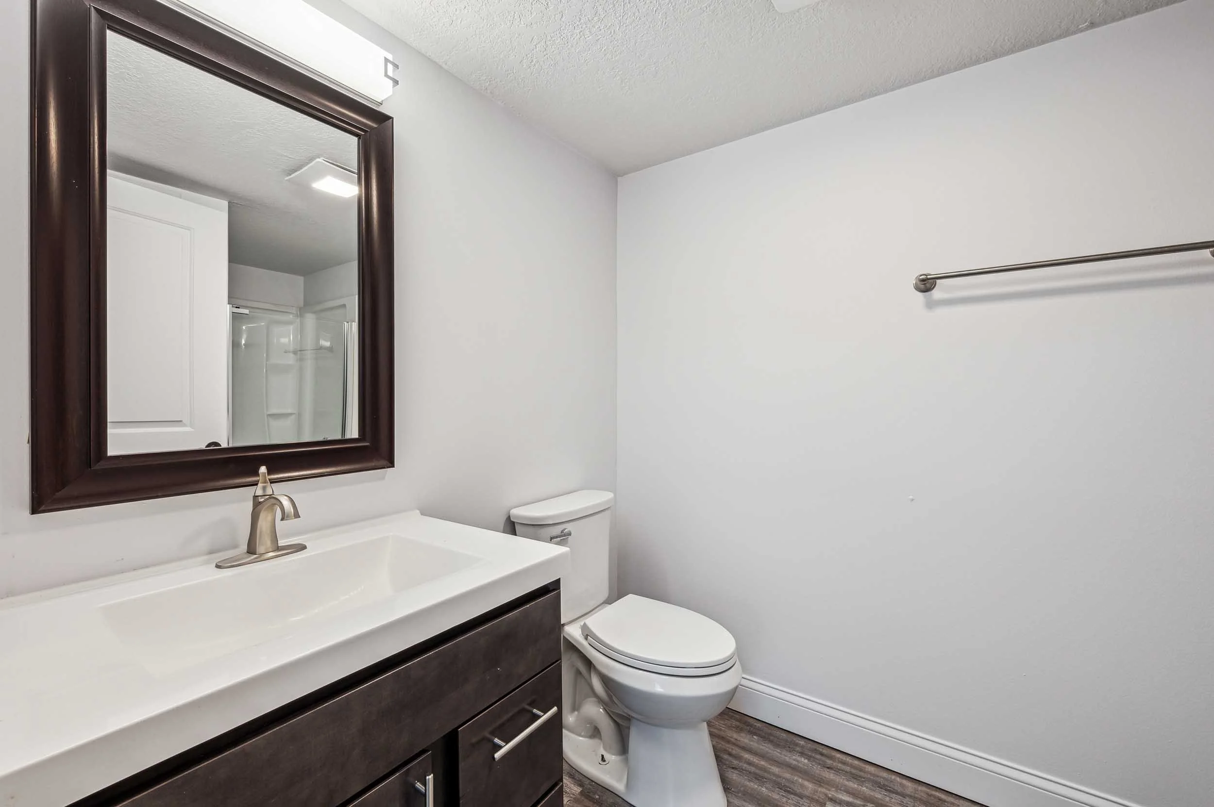 Bathroom with white walls, dark wooden vanity with a white countertop, a mirror with a dark wooden frame, a toilet, a towel bar on the wall, and a shower in the background.