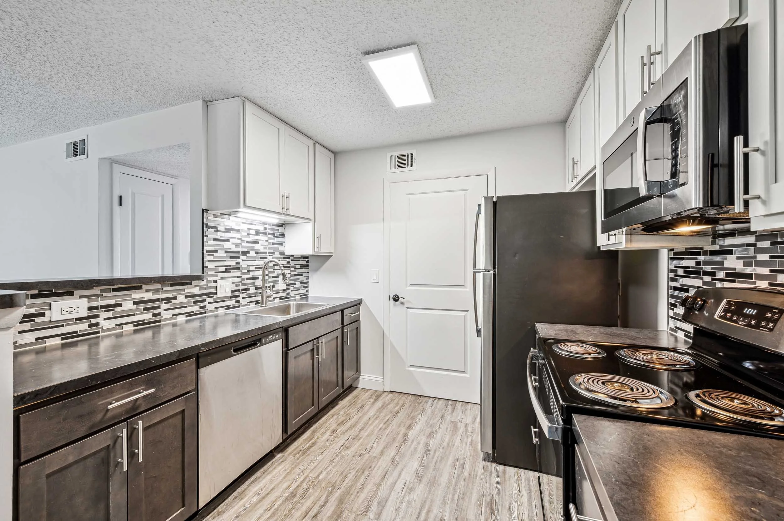 Modern kitchen with white upper cabinets, dark lower cabinets, a stainless steel refrigerator, a microwave, a stove with coil burners, a gray countertop, a mosaic tile backsplash in black, gray, and white, a stainless steel sink, and wood-like floori