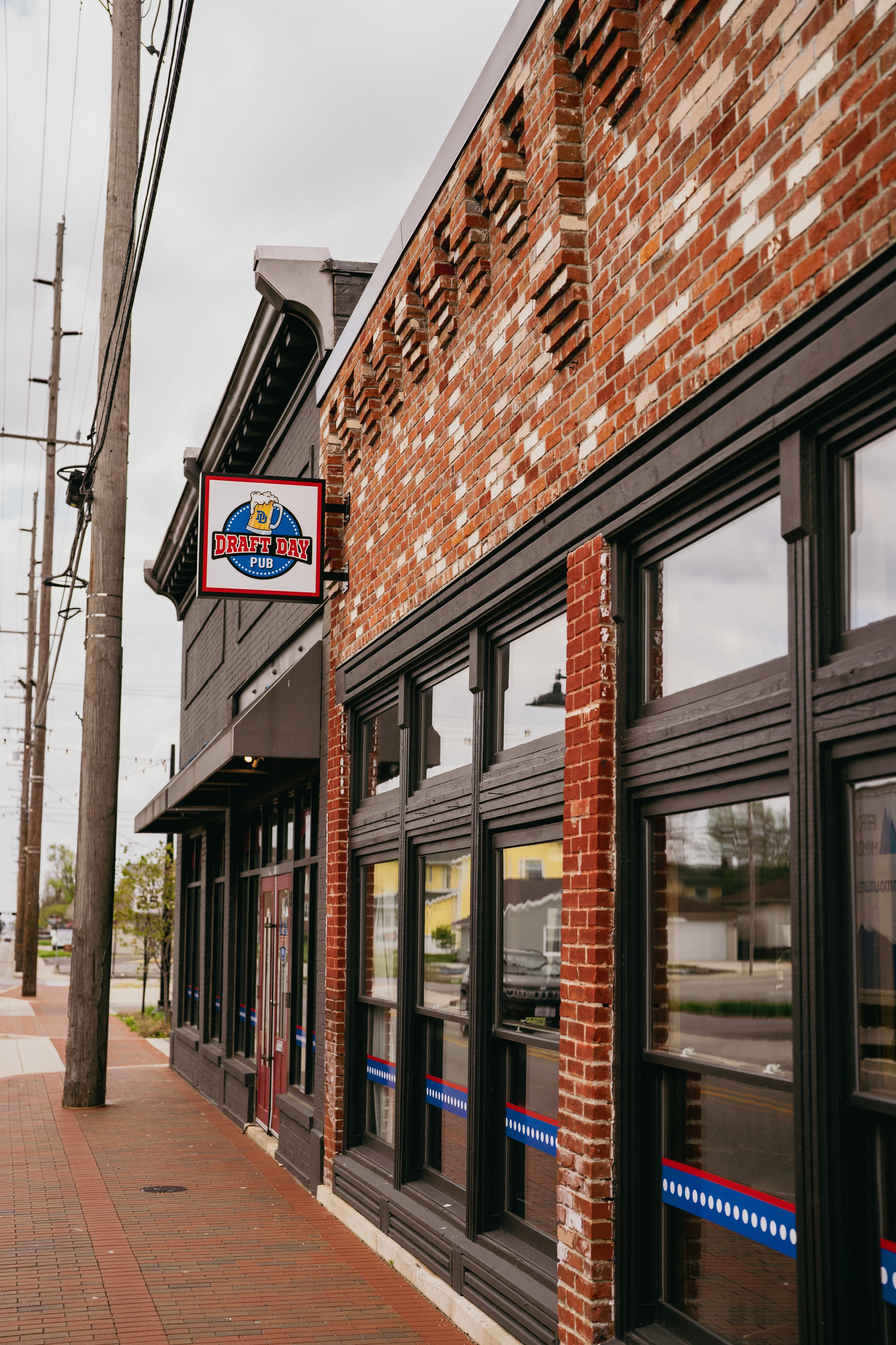 Exterior view of a brick building with black-framed windows and a sign that reads "Draft Day Pub" with an image of a beer mug. There are power lines and a utility pole in the foreground, and the street is paved with bricks.
