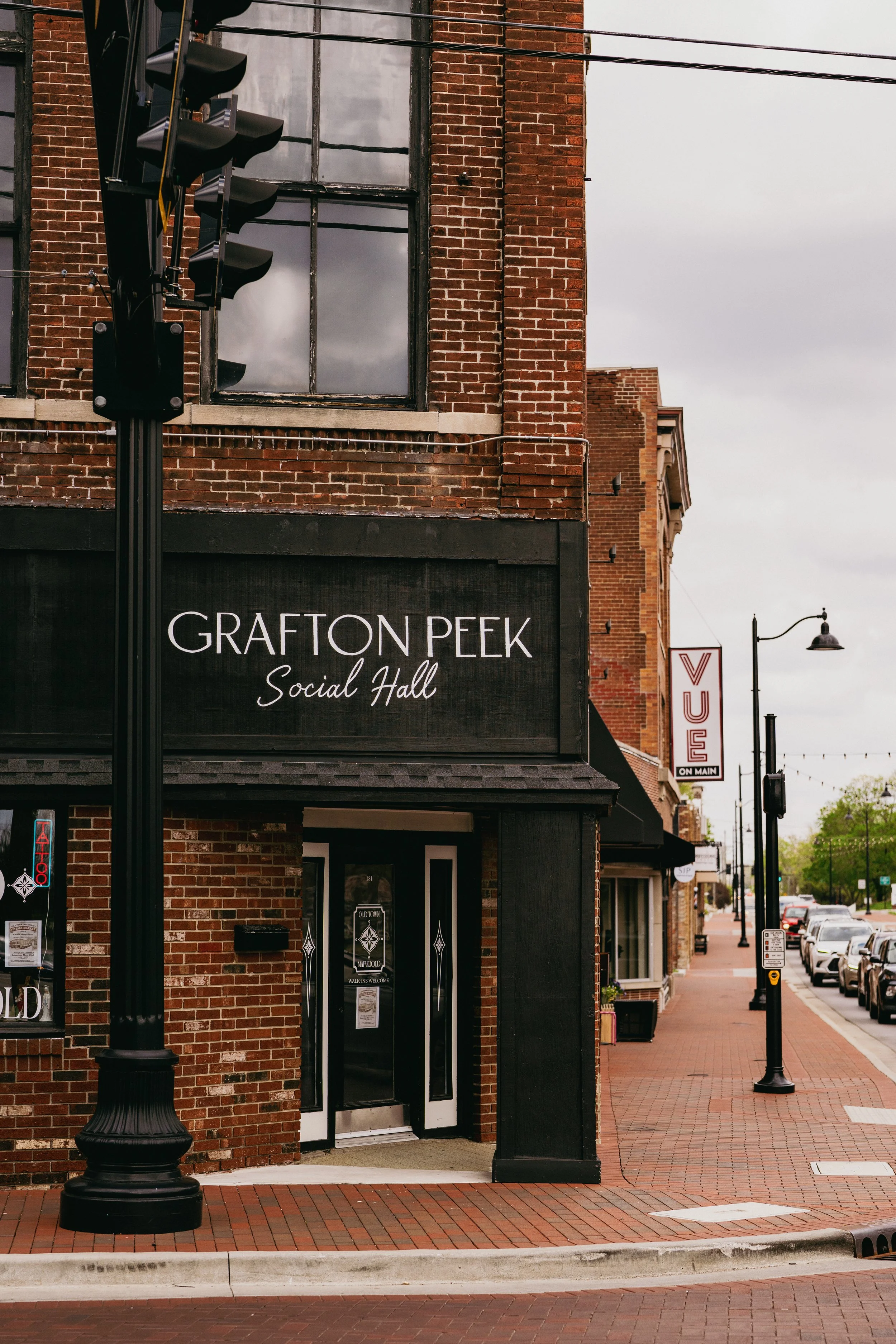 View of a brick building with a black awning that reads 'GRAFTON PEAK Social Hall'. There is a traffic light on a pole in front of the building. The sidewalk is paved with bricks, and there are parked cars and street lamps lining the street, with ove