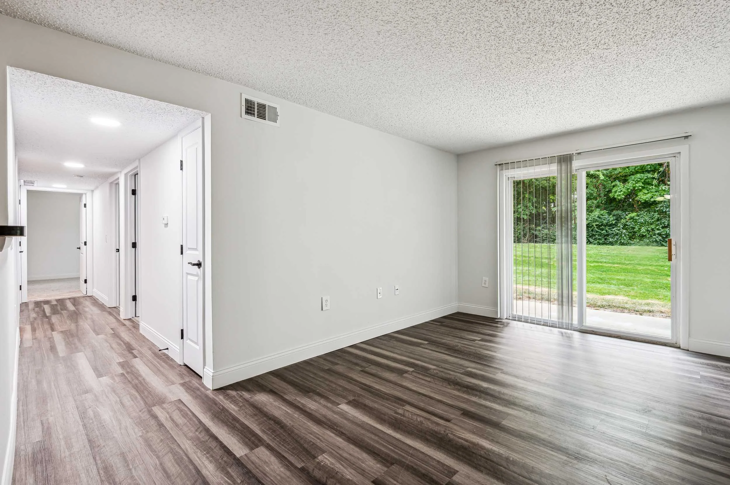 Empty living room with wood flooring, white walls, large sliding glass door, and greenery outside.