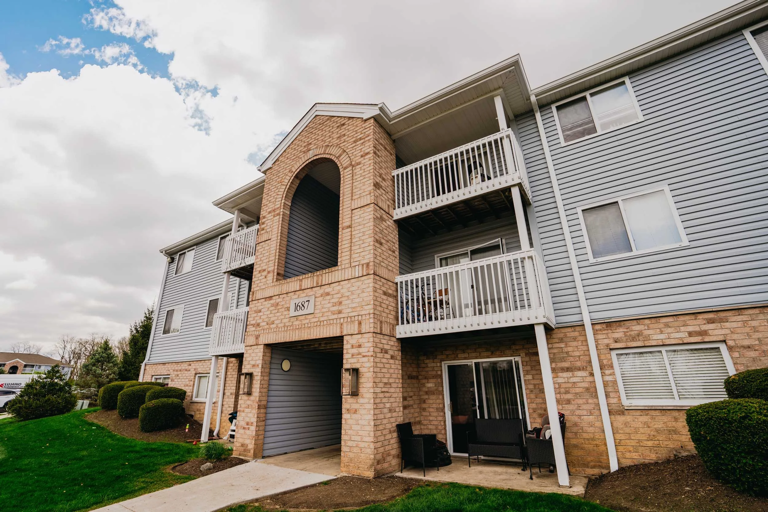 Exterior of a multi-story residential building with brick and siding, featuring balconies with white railings, a garage with a gray door, and well-maintained landscaping including bushes and a lawn under a partly cloudy sky.