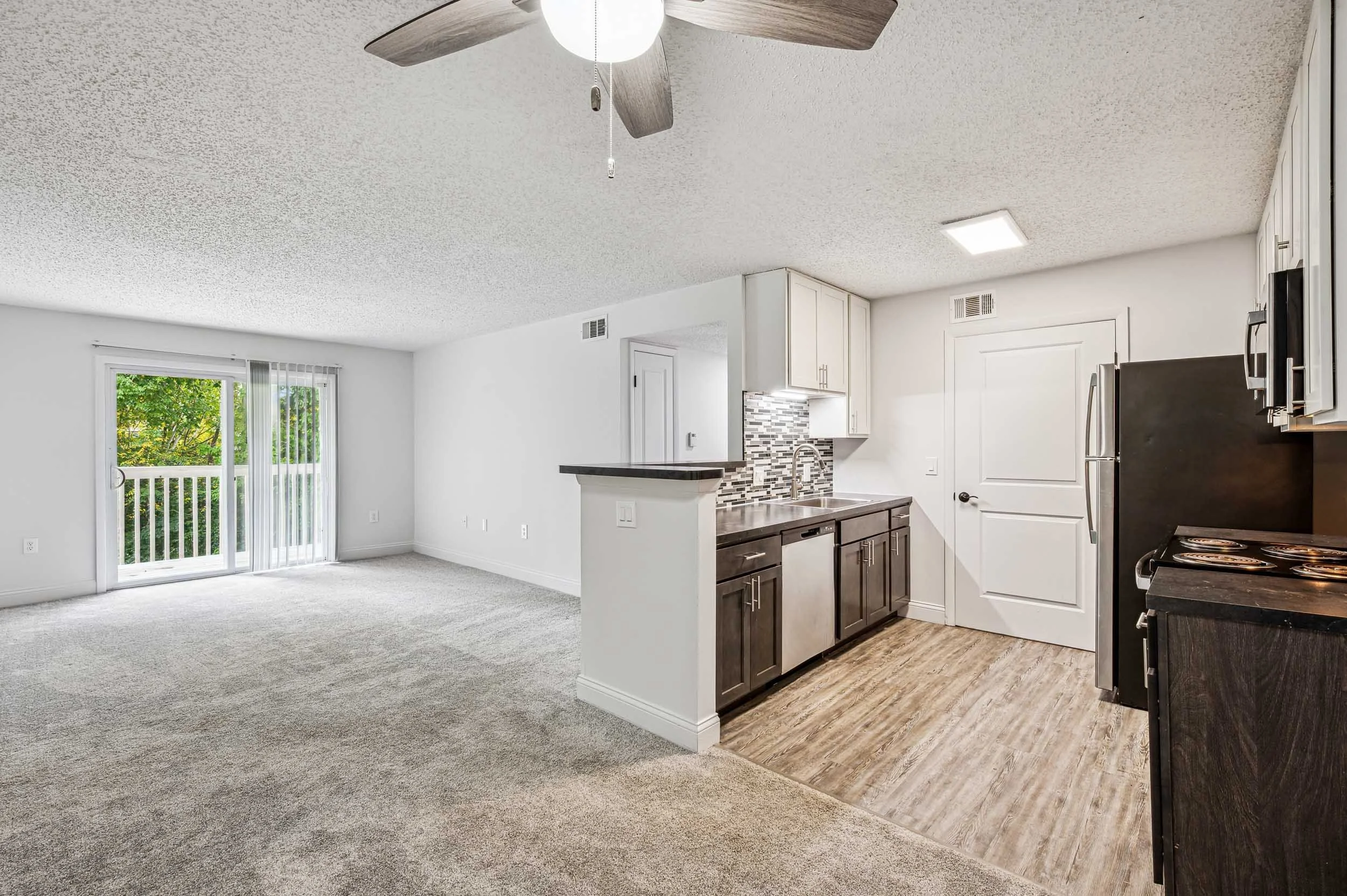 A living space and kitchen area with a sliding glass door opening to a balcony, carpeted living room, white cabinets, a refrigerator, stove, and a small kitchen island with a granite countertop.