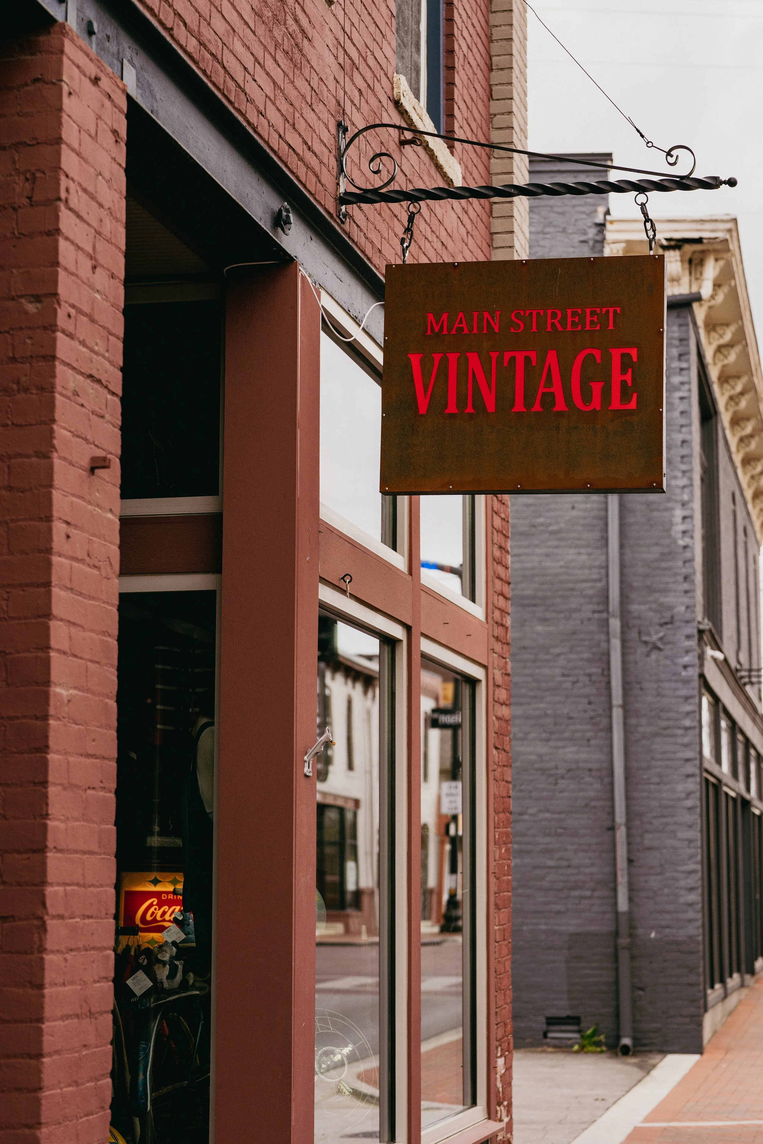 Storefront with a hanging sign reading 'Main Street Vintage' in red letters, brick and siding exteriors, glass windows showing merchandise inside, including Coca-Cola products and stuffed animals, foot traffic on a brick sidewalk.