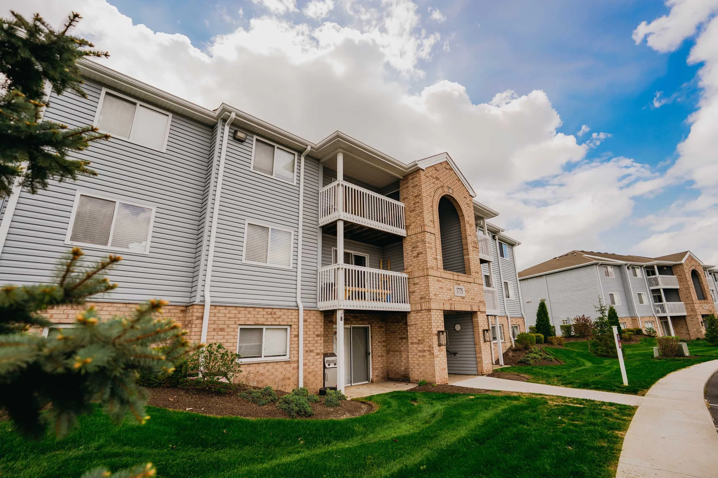 A multi-story apartment building with a brick and gray siding exterior, white balconies, and a sidewalk curving along green landscaped grass and bushes under a partly cloudy blue sky.